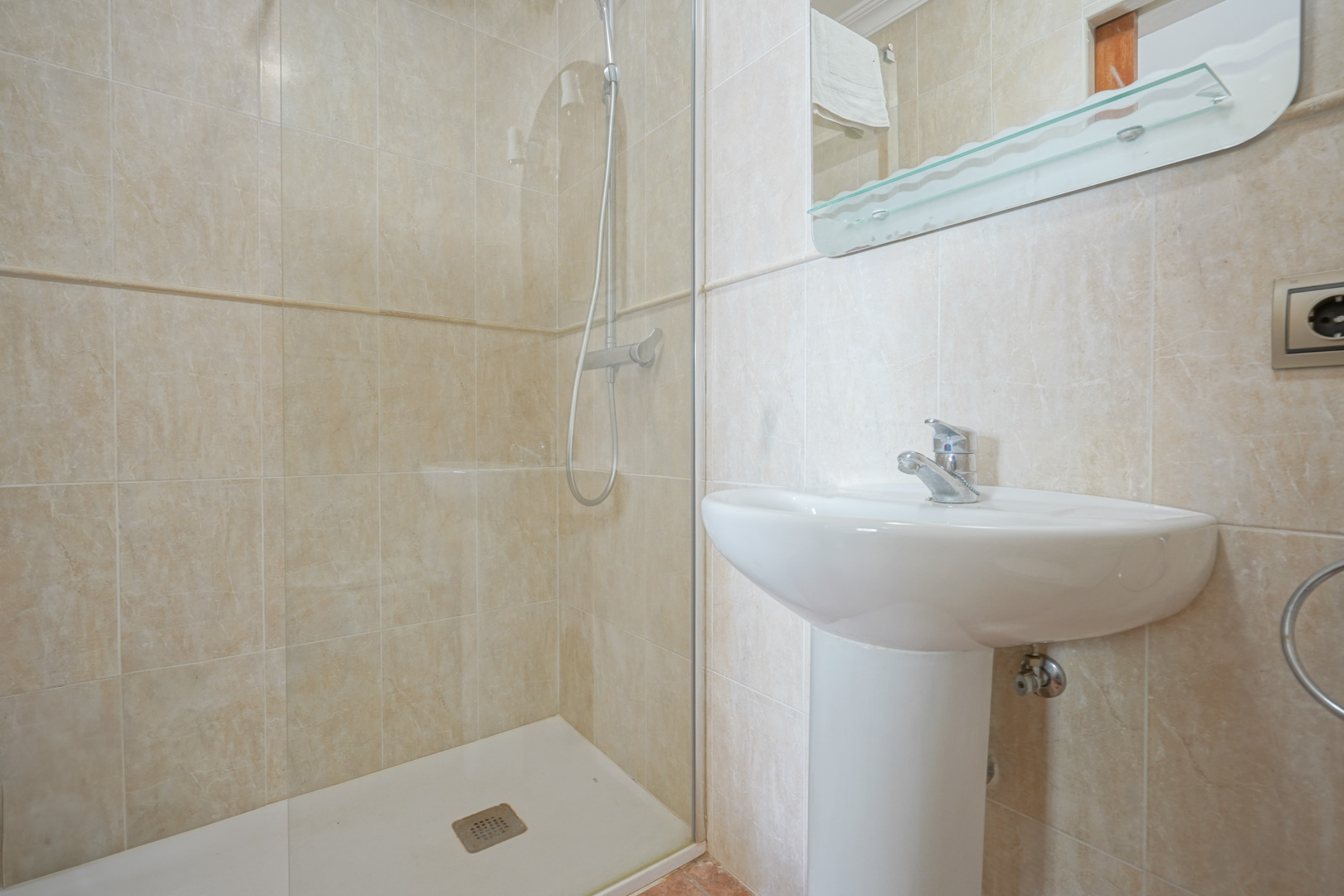 Badezimmer mit beigefarbenen Fliesen, ausgestattet mit einem weißen Säulenwaschbecken und einer ebenerdigen Dusche mit Glastrennwand und Chromarmaturen.