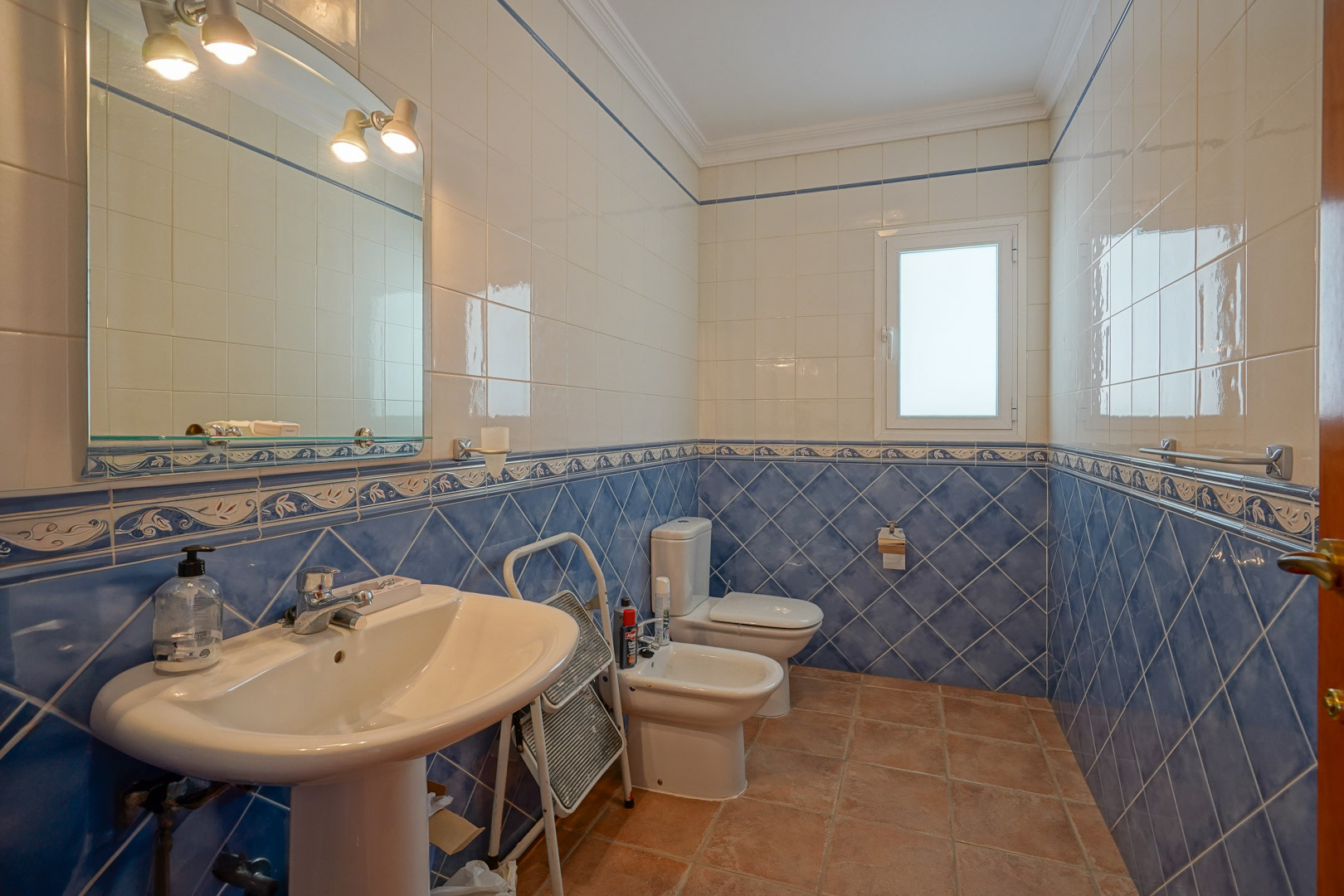 Badezimmer mit blau-weißen Wandfliesen, Terrakottaboden und klassischen weißen Sanitäranlagen wie Standwaschbecken und Bidet unter einem beleuchteten Spiegel.