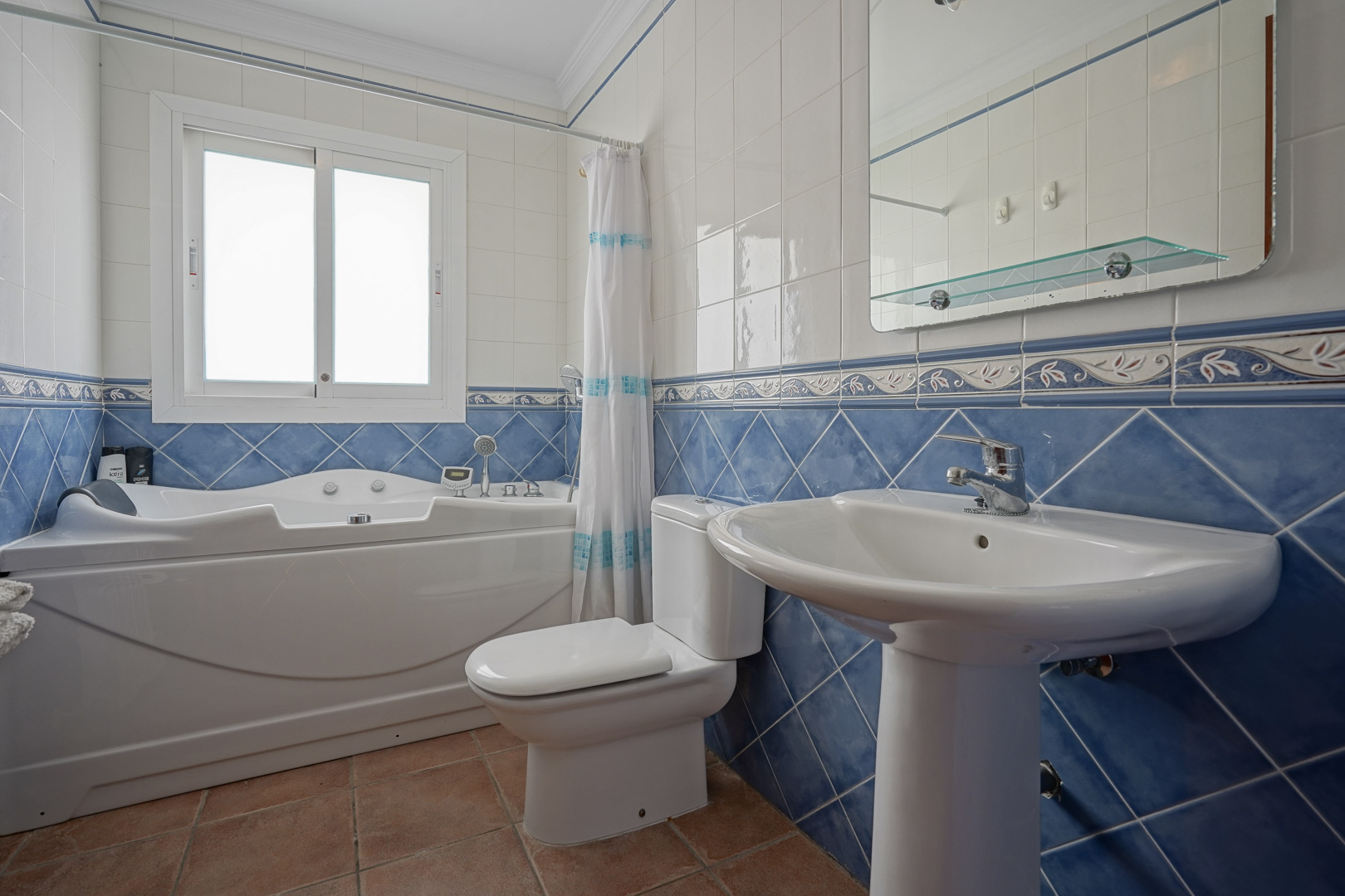 Badezimmer mit Whirlpool-Badewanne, Standwaschbecken und blau-weißen Wandfliesen. Ausgestattet mit Terrakottaboden und einem Fenster für Tageslicht.