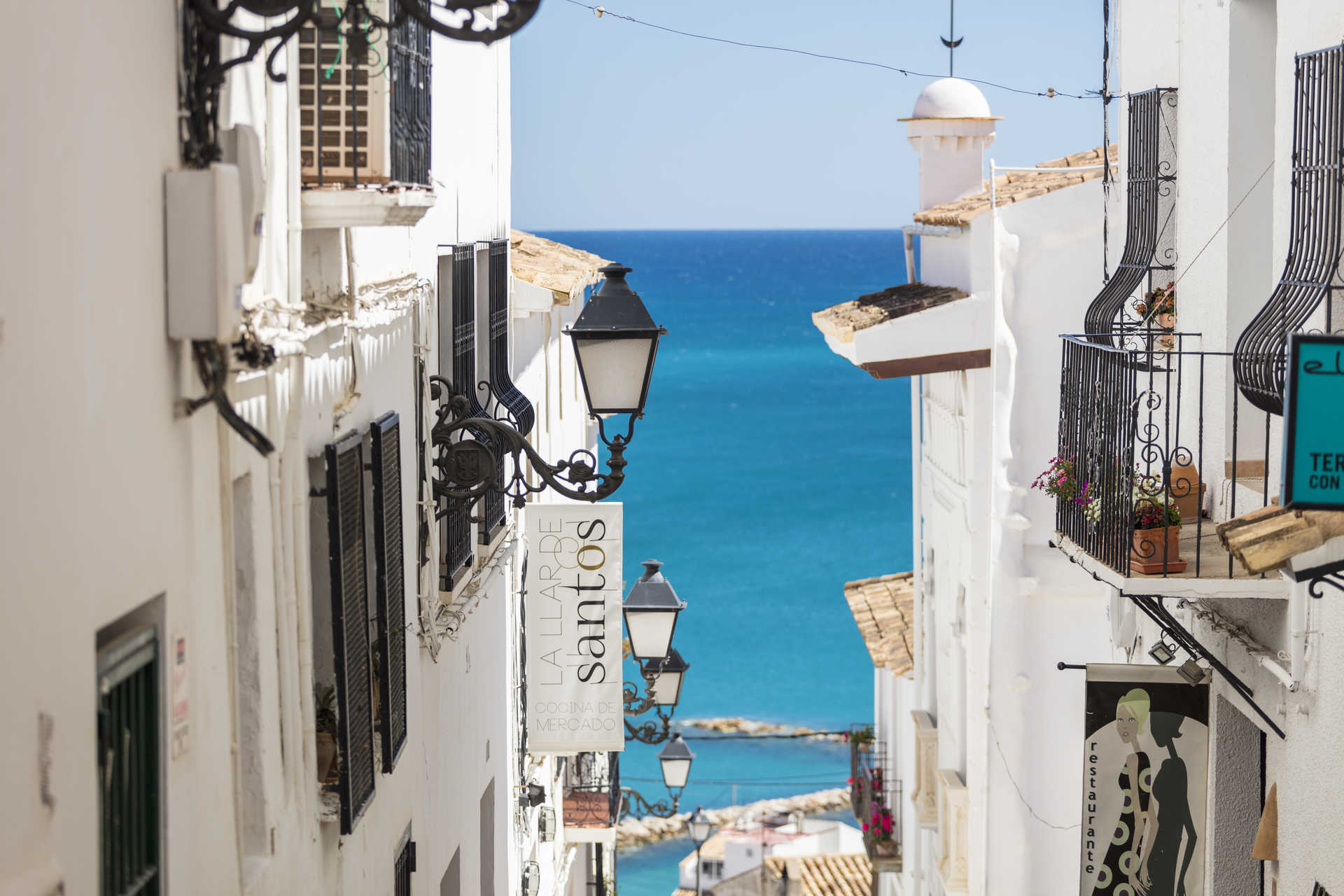 Blick in eine schmale Gasse der Altstadt von Altea mit weißen Fassaden, schmiedeeisernen Laternen und freiem Blick auf das türkisfarbene Mittelmeer.