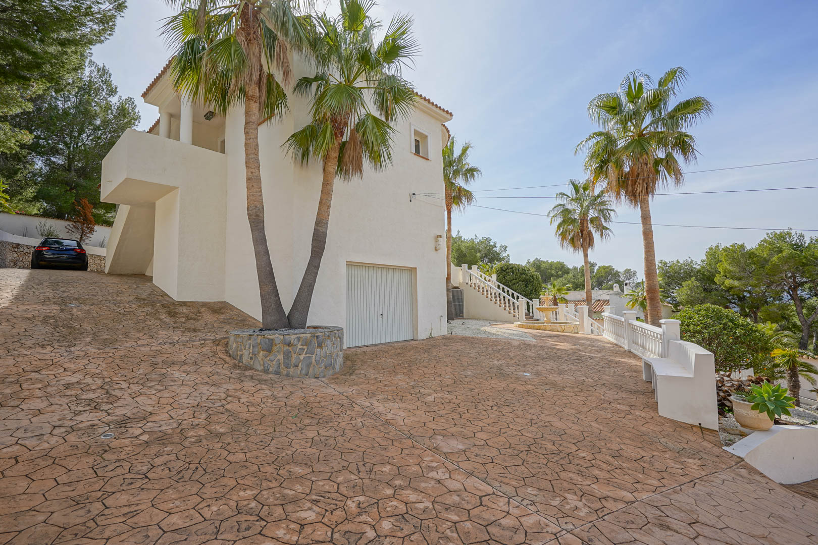 Gepflasterte Einfahrt mit gemustertem Beton führt zur Garage und zum Eingang. Hohe Palmen und die weiße Fassade prägen den mediterranen Außenbereich der Villa.