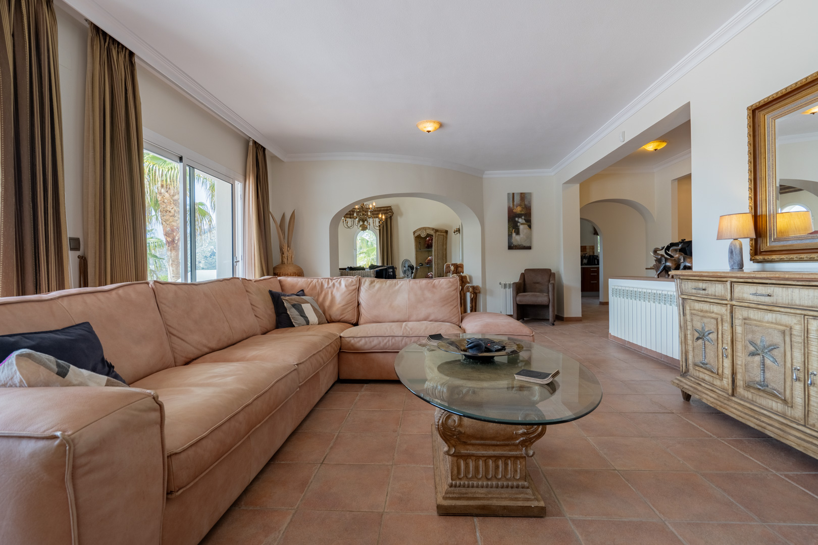 Großzügiges Wohnzimmer mit Terrakottaböden, großem Ecksofa und Rundbogen-Durchgängen. Ein Glastisch mit Steinsockel und ein geschnitztes Sideboard zieren den Raum.