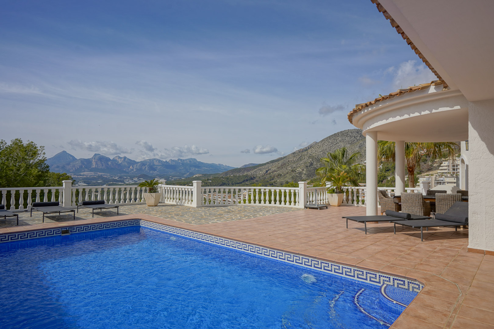 Hauptterrasse mit blau gefliestem Pool, Sonnenliegen und überdachter Veranda. Sie bietet einen Panoramablick auf die Berge Puig Campana und Sierra Aitana.