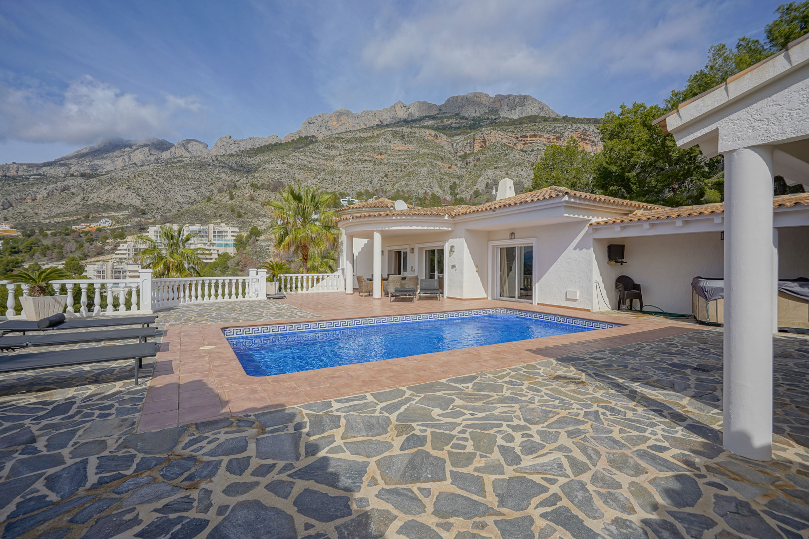 Hauptterrasse mit rechteckigem Pool, Mosaikbordüre und Blick auf die Sierra de Bernia. Der Außenbereich ist mit Natursteinpflaster, Terrakottafliesen und einer weißen Balustrade gestaltet.