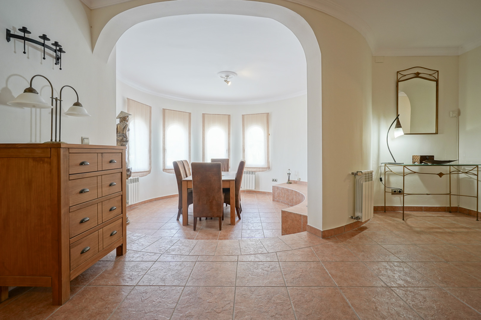 Heller Essbereich mit Rundbogenfenstern und Terrakottaboden. Die kreisförmige Architektur bietet Platz für eine Holz-Sitzgruppe und geht fließend in den Flur über.