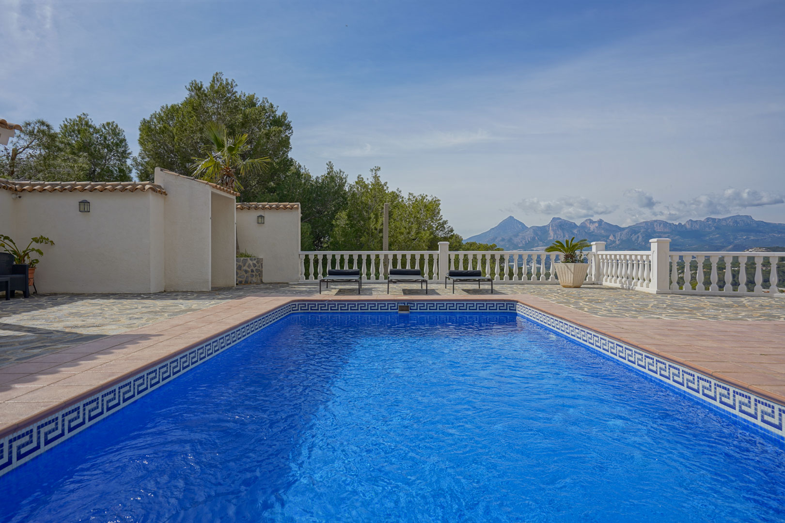Poolterrasse mit Natursteinpflaster und weißer Balustrade, die einen weiten Blick auf die Gebirgsketten des Puig Campana und der Sierra Aitana bietet.