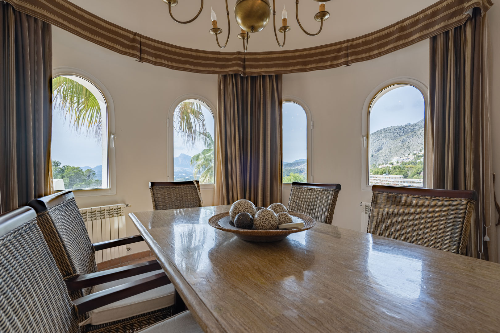 Rundes Esszimmer mit poliertem Steintisch und Rattanstühlen. Vier Rundbogenfenster bieten einen Panoramablick auf die Sierra de Bernia und die umliegende Berglandschaft.