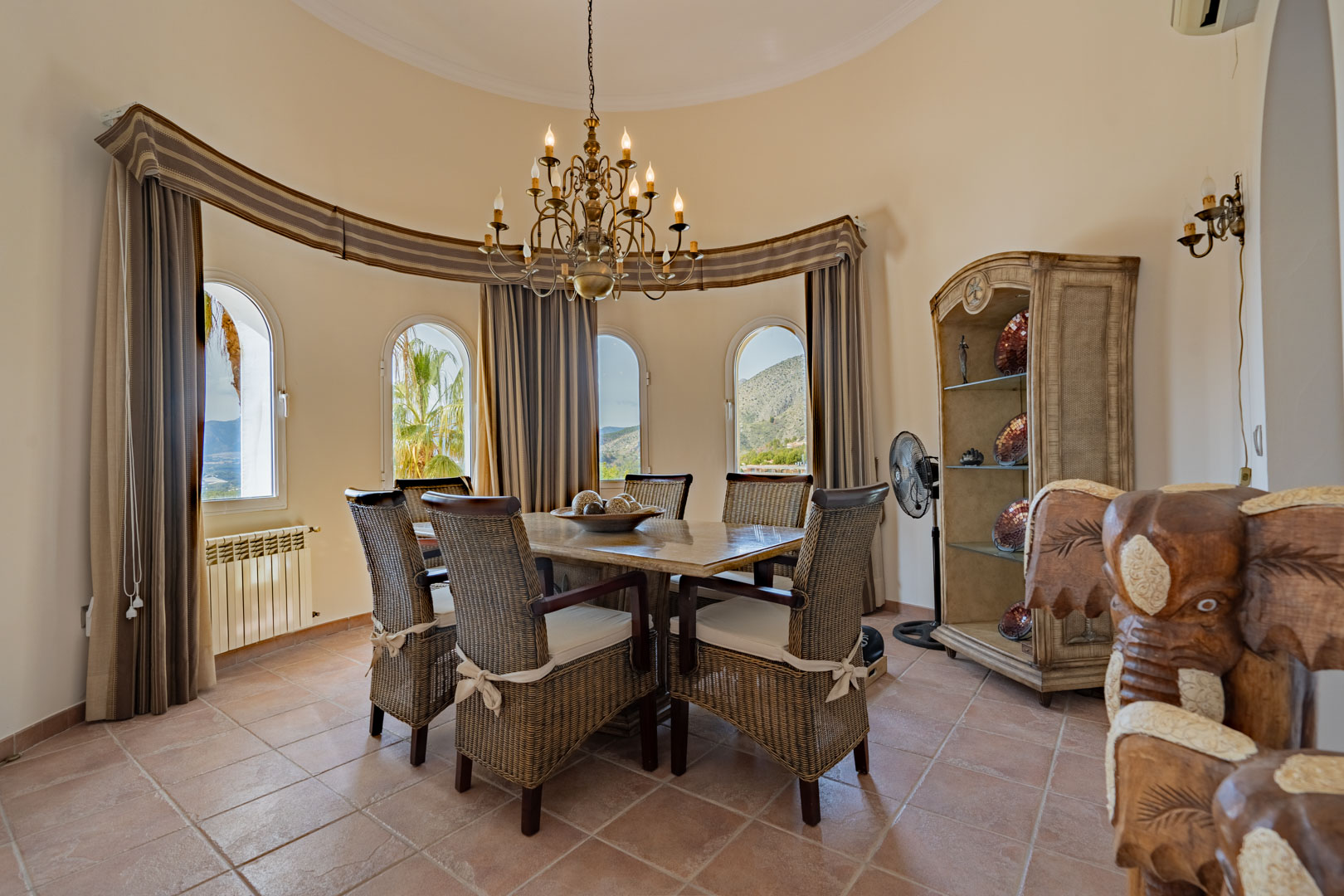 Rundes Esszimmer mit Steintisch, Korbstühlen und klassischem Kronleuchter. Vier Rundbogenfenster bieten einen Panoramablick auf das Sierra de Bernia-Gebirge.
