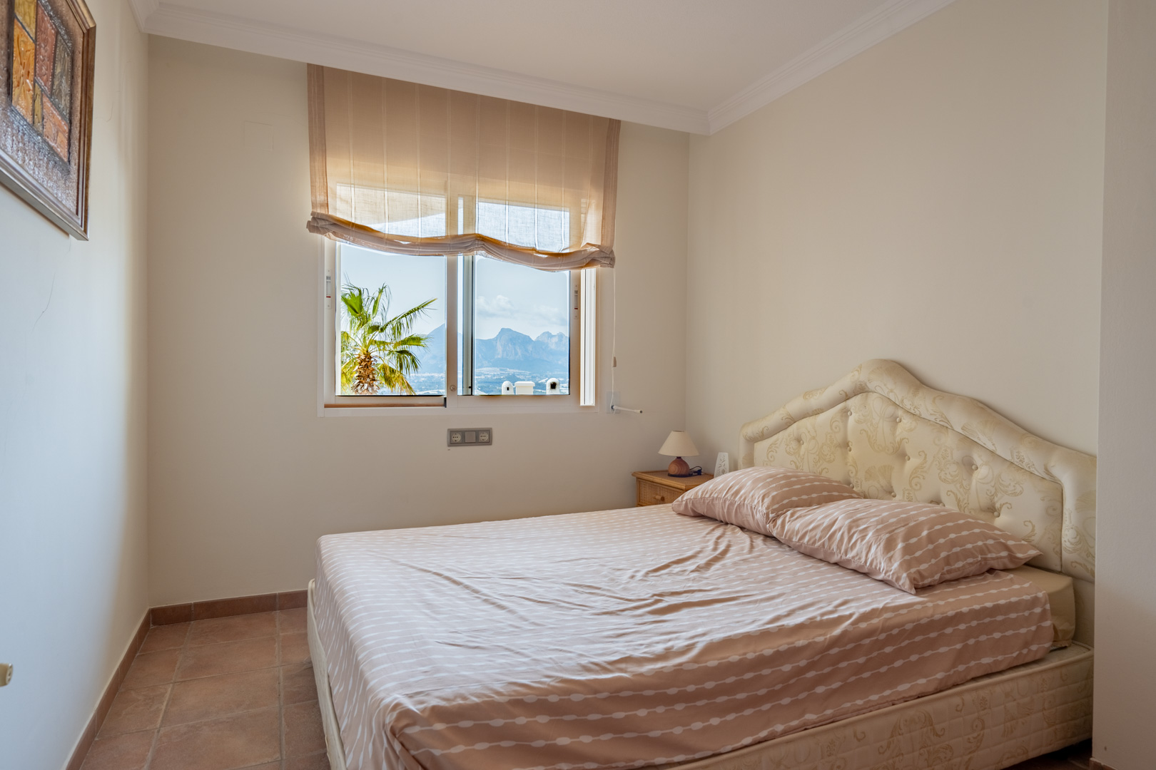 Schlafzimmer mit Fliesenboden und gepolstertem Kopfteil. Das Fenster bietet einen Blick auf die Bergkette und eine Palme im Außenbereich.