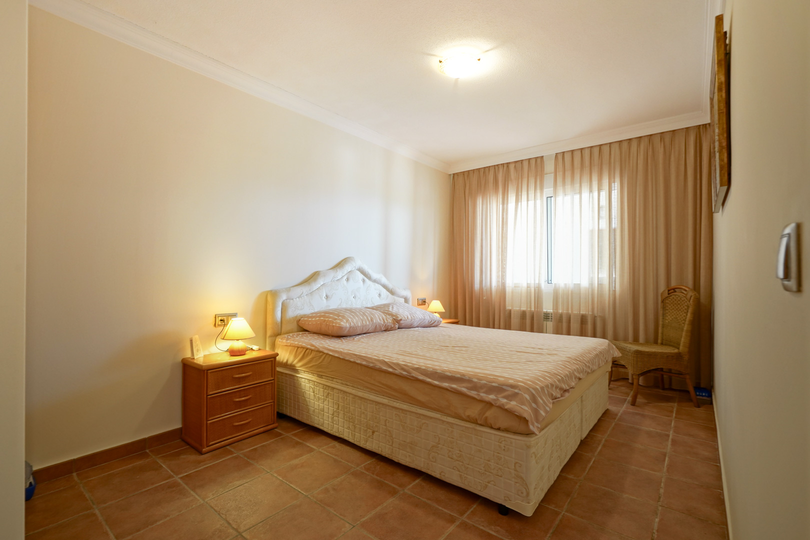 Schlafzimmer mit Terrakotta-Fliesenboden, Doppelbett mit gepolstertem Kopfteil, Nachttisch aus Holz und Fenster mit lichtdurchlässigen Vorhängen.