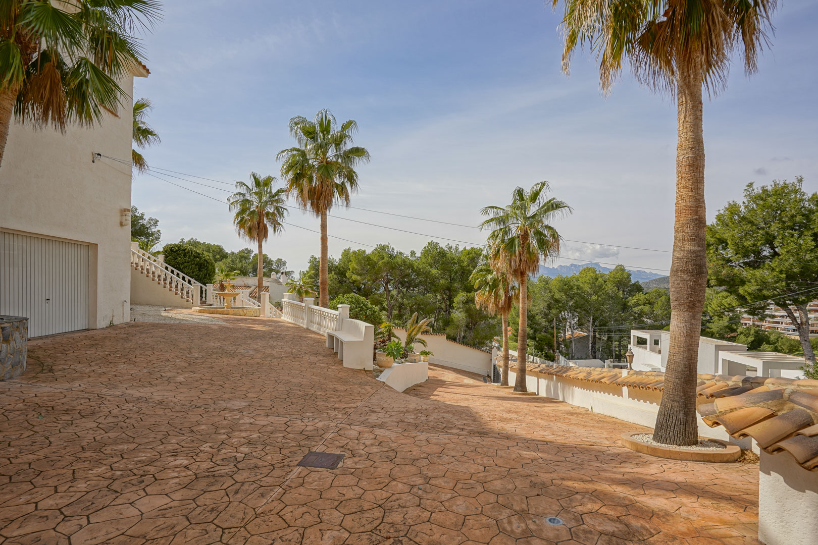 Villeneingang mit großzügiger Einfahrt aus geprägtem Beton, hohen Palmen und Bergblick. Der Bereich umfasst einen Garagenzugang und klassische weiße Balustraden.