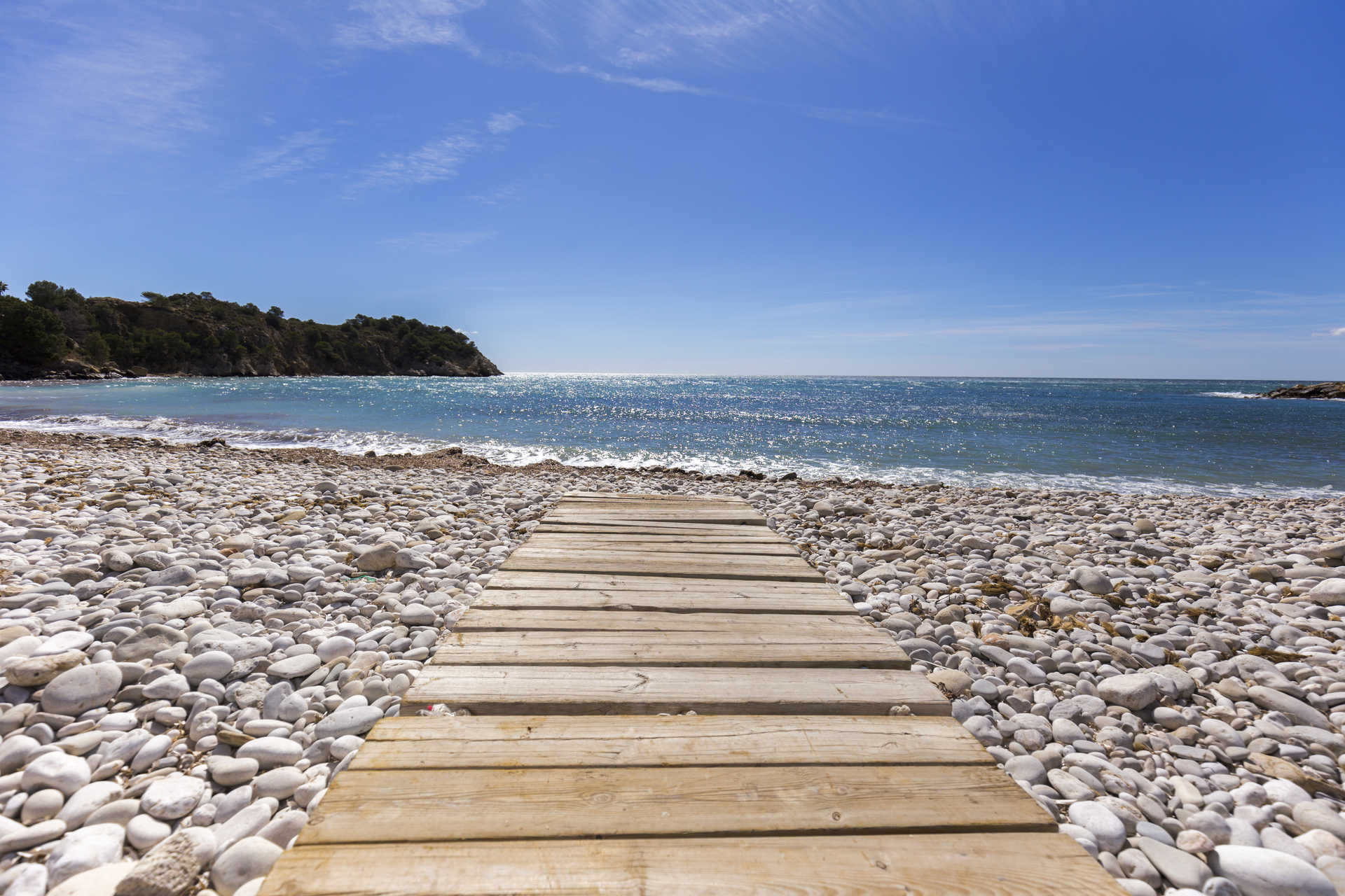 Weißer Kieselstrand in Altea mit einem Holzsteg, der zum türkisfarbenen Mittelmeer führt, umgeben von einer natürlichen Küstenlandschaft.