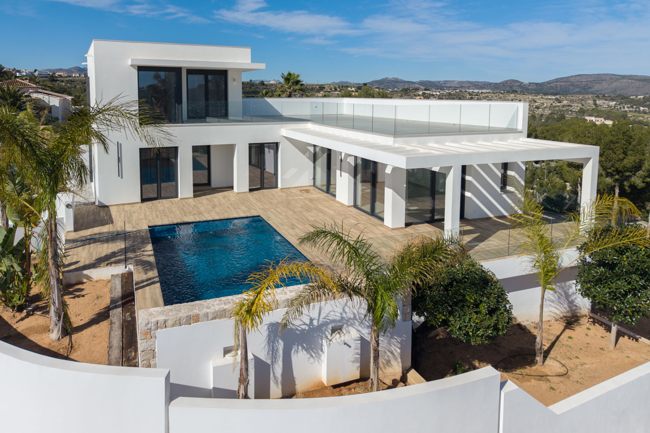Luftaufnahme der modernen Villa mit blauem Pool, Holzterrasse und Glasgeländer in Benimeit, Moraira.