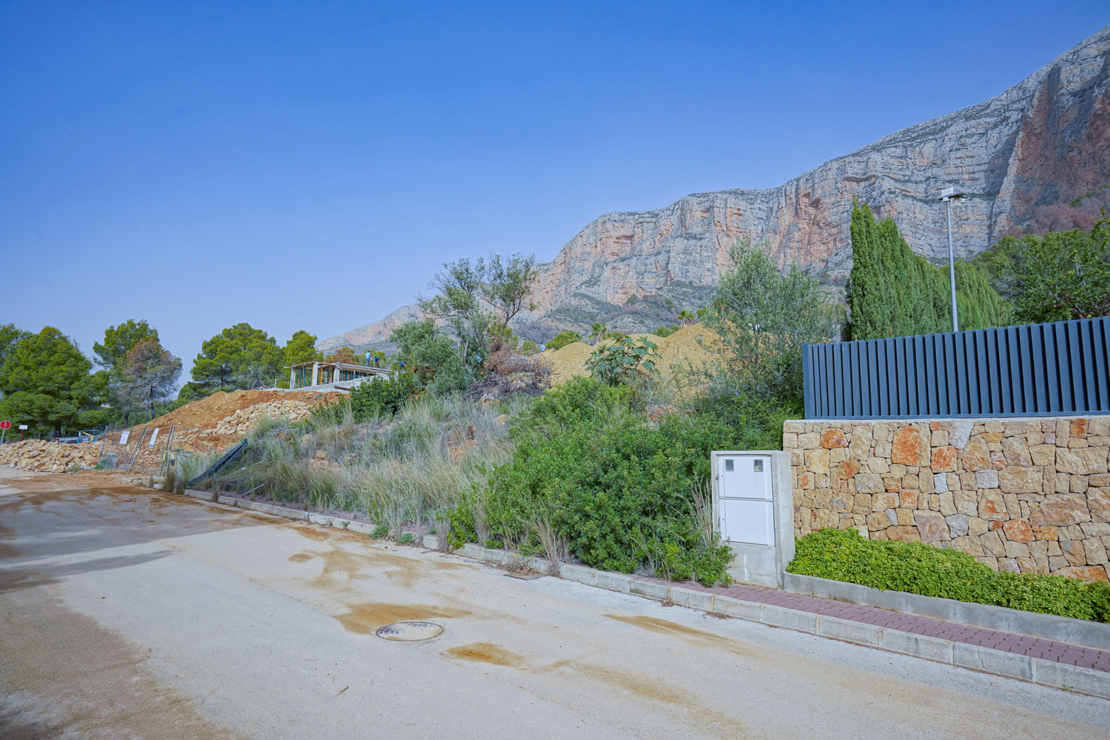 Baustelle in der Rohbauphase am Fuße des Montgó-Massivs, mit Blick auf die markanten Felswände und die bereits errichtete Natursteinmauer.