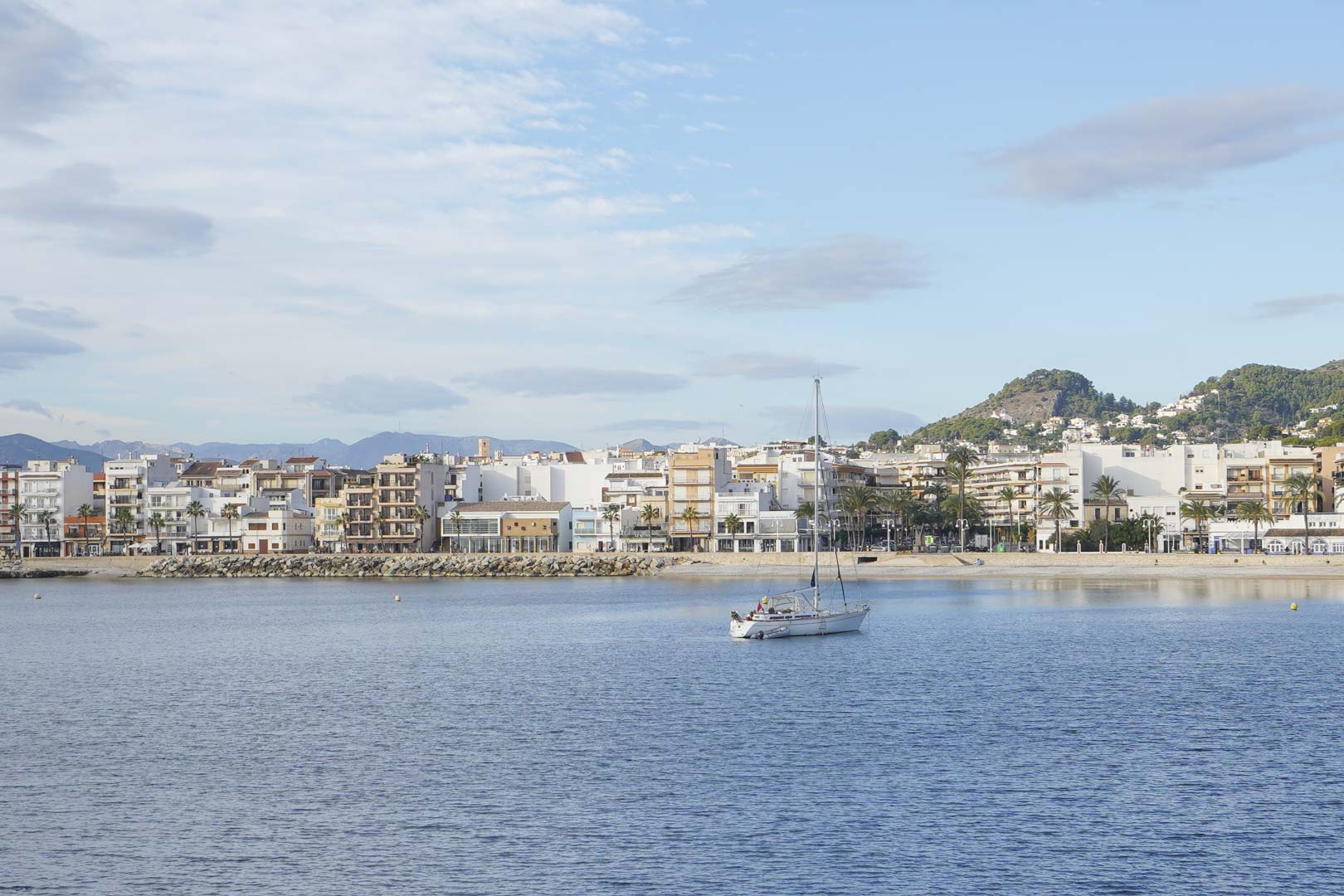 Panoramablick auf die Küste von Jávea mit einem Segelboot auf ruhigem Wasser, vor der Kulisse mediterraner Gebäude und ferner Berge.