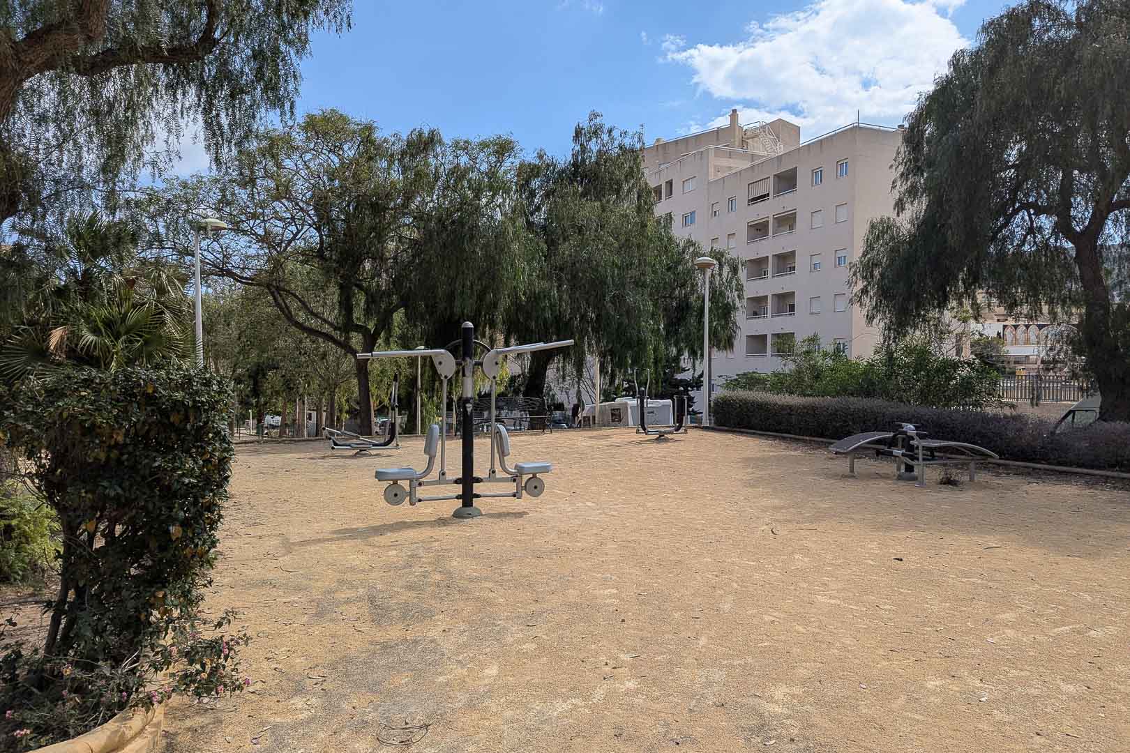 Fitnessbereich im Freien mit verschiedenen Trainingsgeräten, eingebettet in eine parkähnliche Anlage mit altem Baumbestand und Wohngebäuden.
