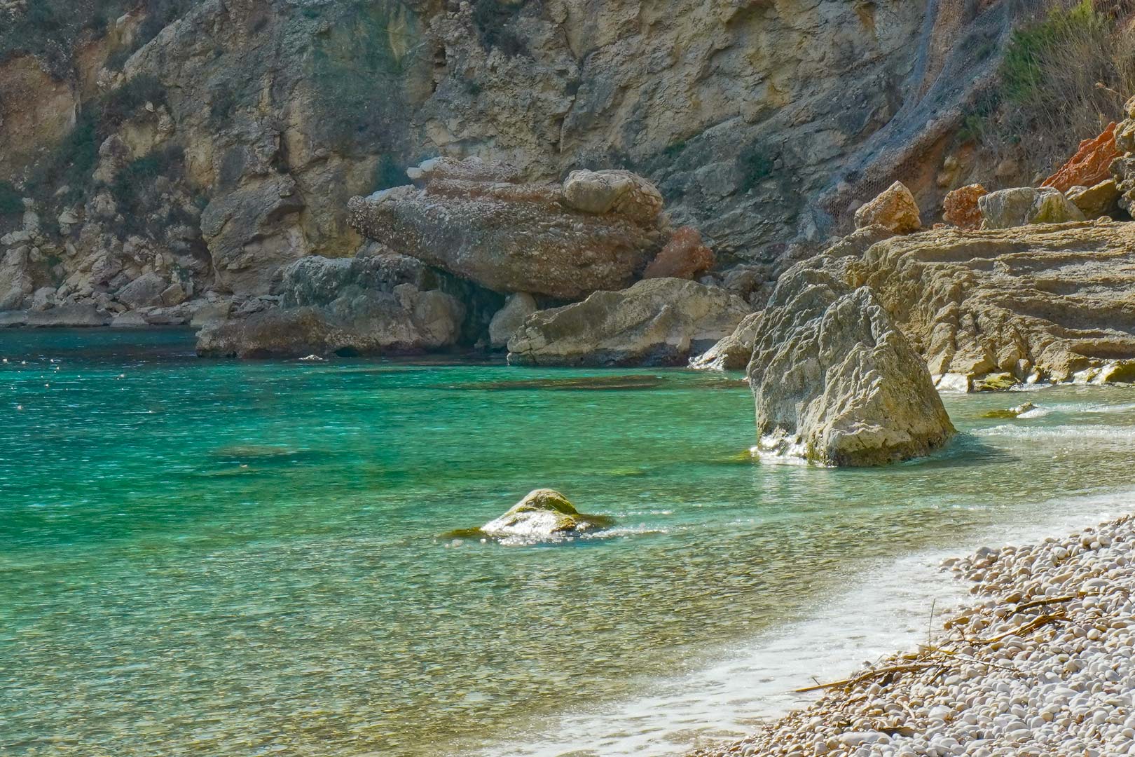 Blick auf eine nahegelegene natürliche Bucht mit kristallklarem, türkisblauem Wasser, weißem Kieselstrand und markanten Kalksteinfelsen.