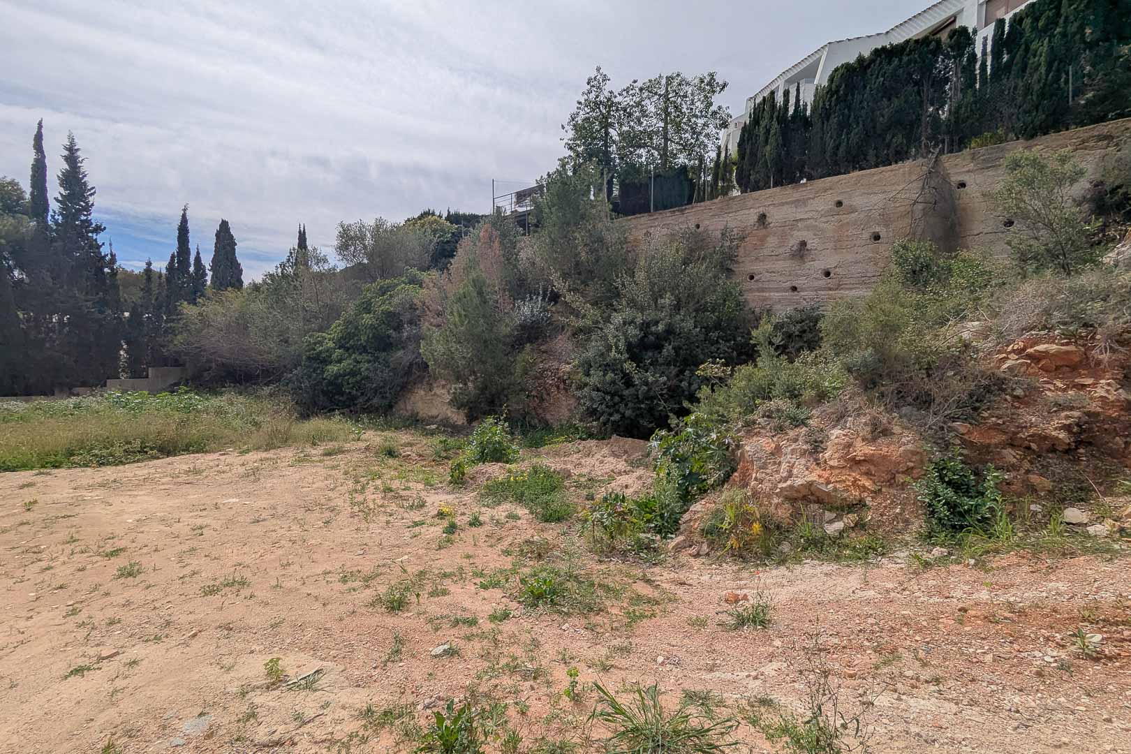 Erhöhtes Grundstück in La Corona mit Betonstützmauer und mediterraner Vegetation. Das Gelände ist bereit für den Bau eines maßgeschneiderten Villenprojekts.