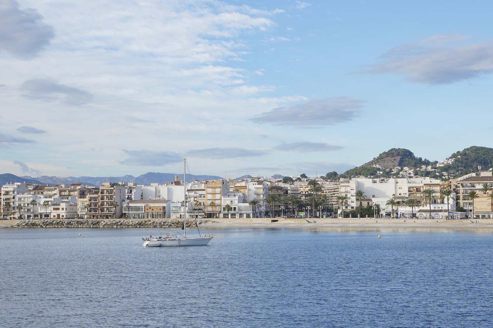 Panoramablick auf den Hafen von Jávea mit einem Segelboot in der Bucht, der Uferpromenade und den charakteristischen Bergen im Hintergrund.