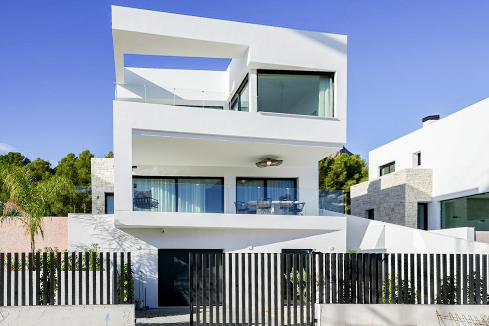 Außenansicht einer modernen dreistöckigen Villa mit Balkonen, Glasgeländern und einem schwarzen Lattenzaun.