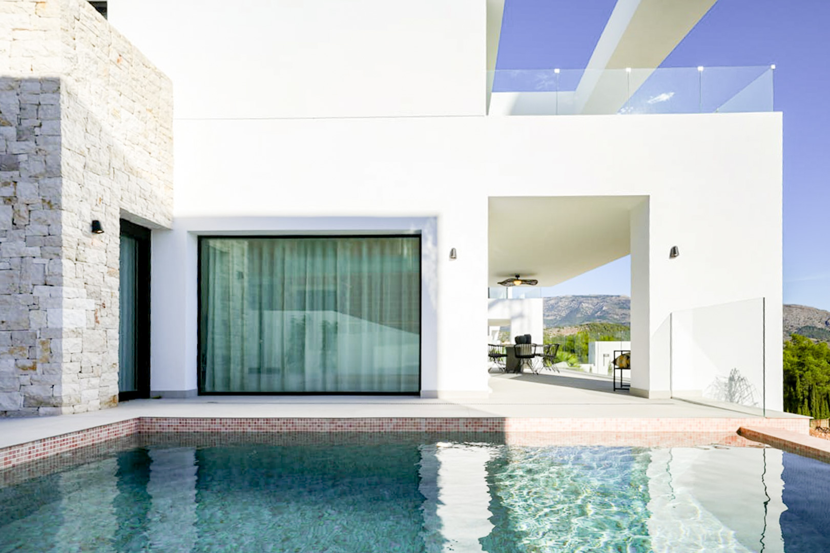 Blick auf den Pool mit Mosaikfliesenrand, Steinfassade und überdachter Terrasse mit Essbereich im Freien.