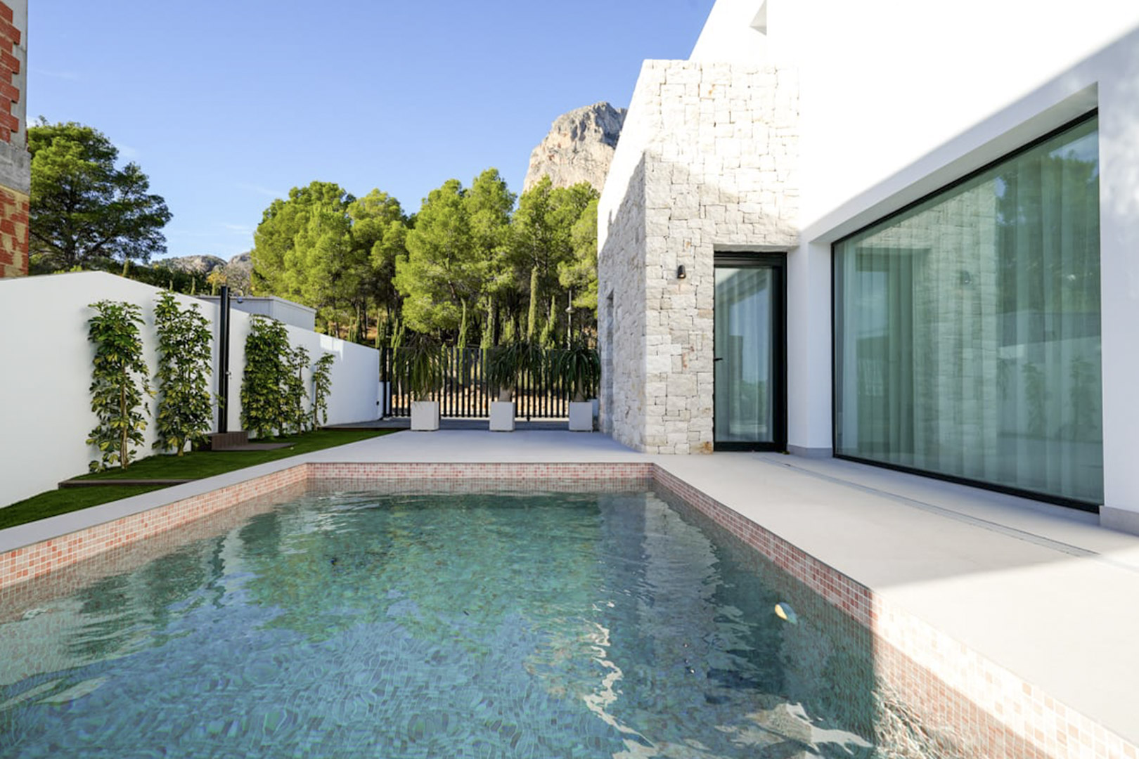 Blick auf den privaten Pool mit Mosaikrand und weißer Steinfassade, umgeben von üppigem Grün.