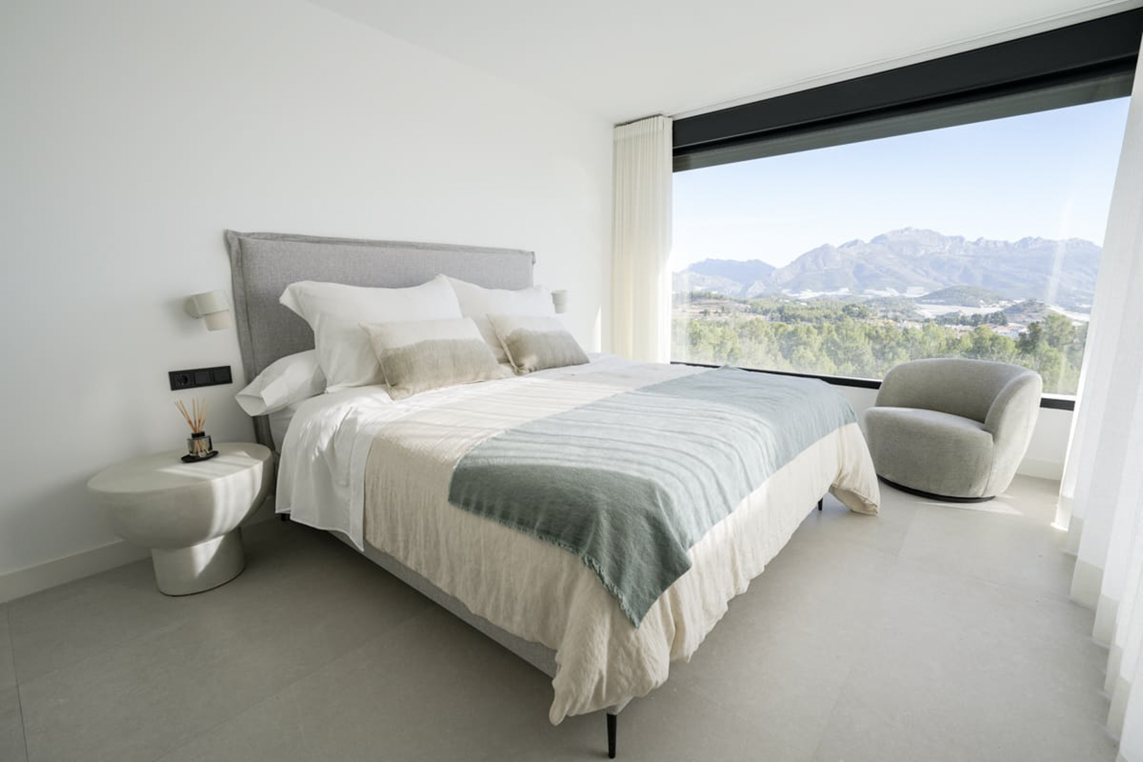 Helles Schlafzimmer mit Doppelbett, grauem Sessel und Bergblick durch ein großes Fenster.