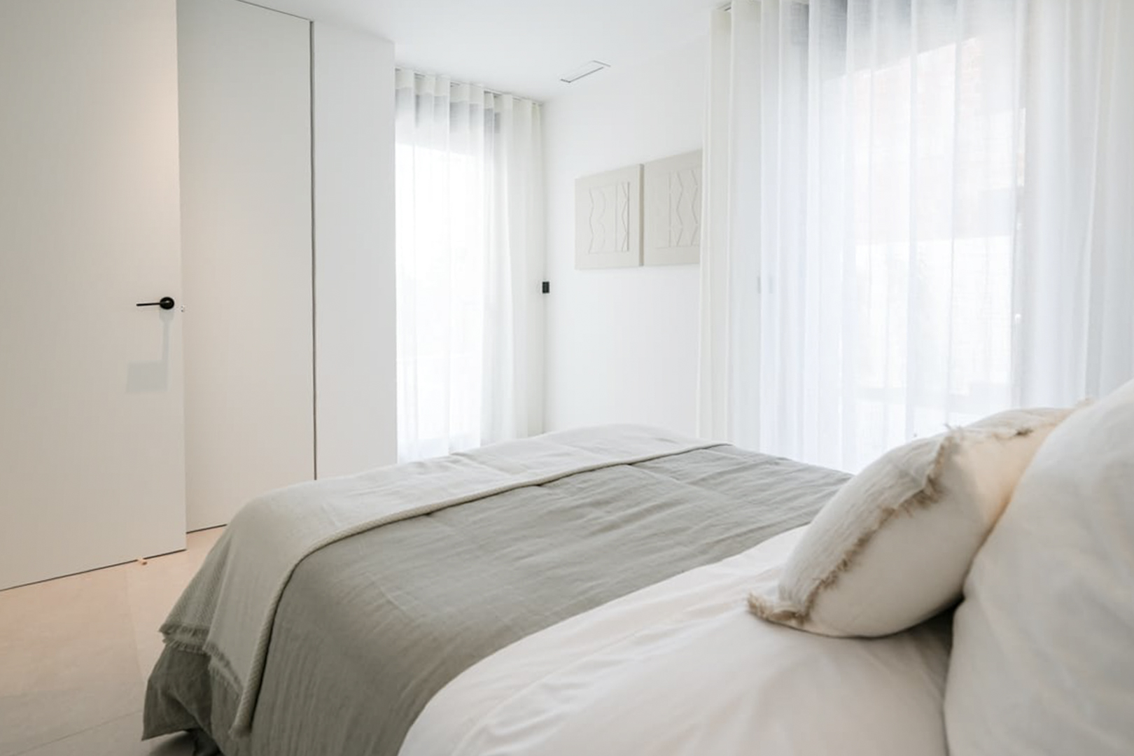 Helles Schlafzimmer mit grauer und weißer Bettwäsche, transparenten Vorhängen und minimalistischer Wanddekoration.