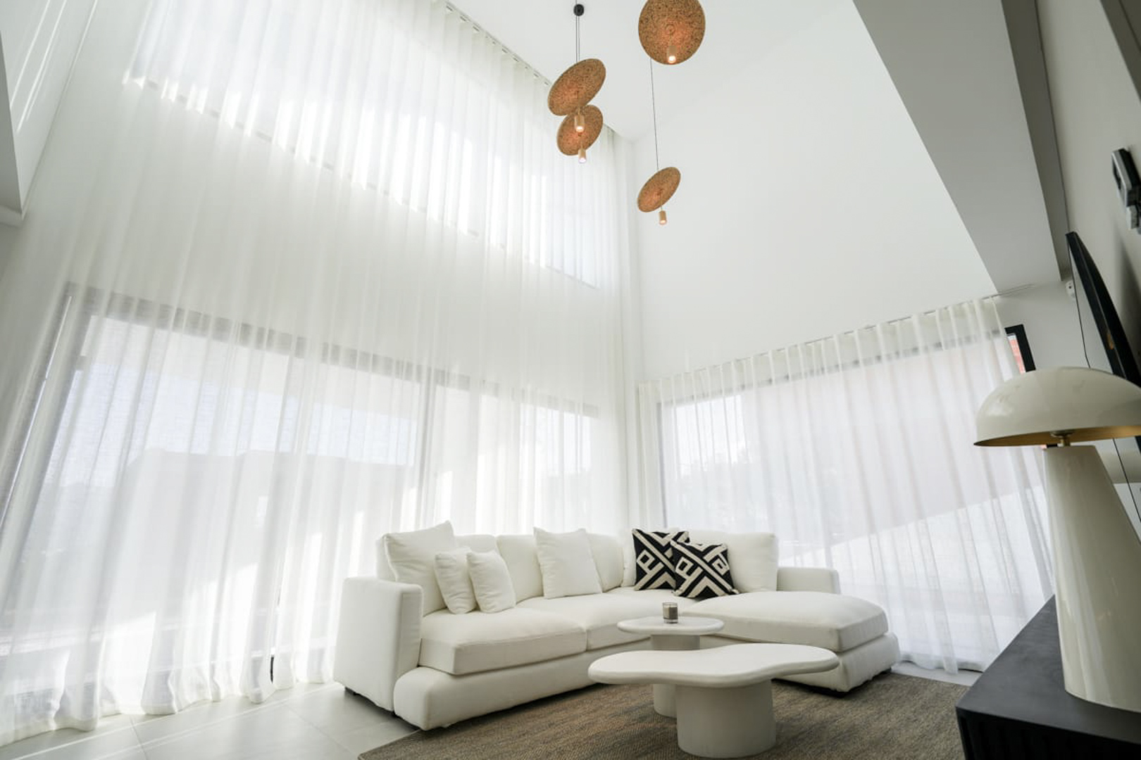 Helles Wohnzimmer mit weißen Vorhängen, Ecksofa und Design-Pendelleuchten, die ein modernes, minimalistisches Ambiente schaffen.