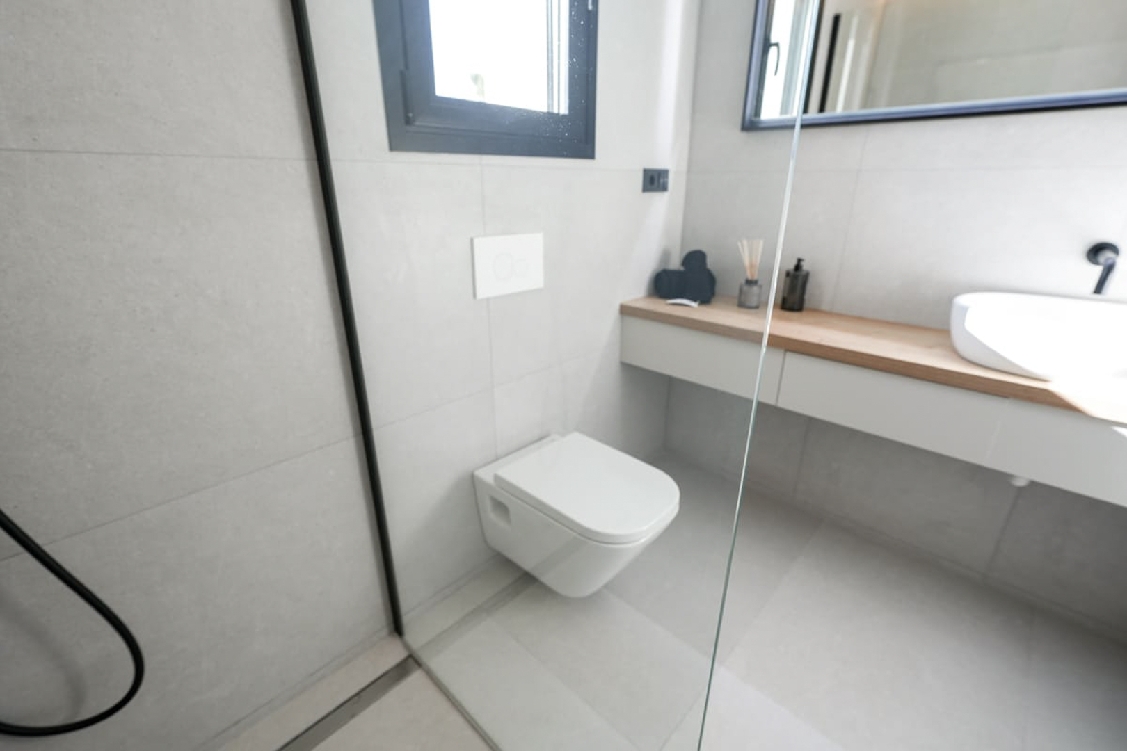 Modernes Badezimmerdetail mit grauen Fliesen, wandhängendem WC und heller Holzablage.