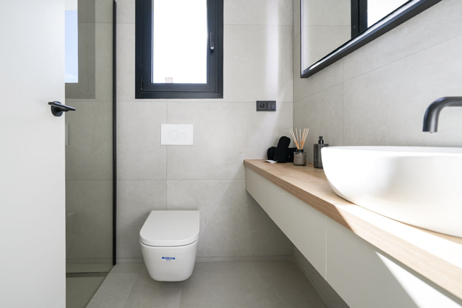 Modernes Badezimmerdetail mit heller Holzablage, weißem Waschbecken und neutralen grauen Fliesen.