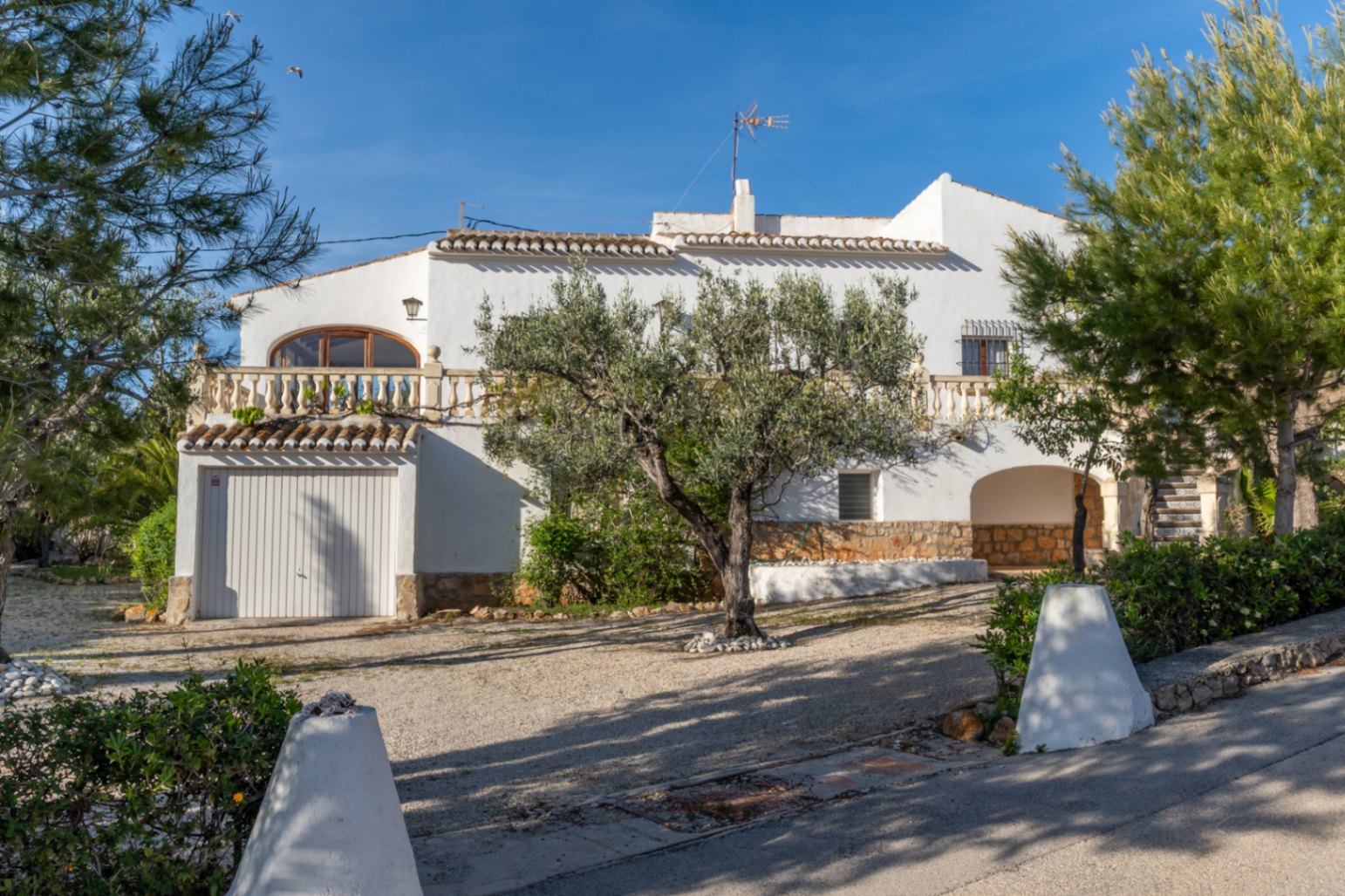 Außenansicht der Villa mit traditioneller Architektur, integrierter Garage, Balustradenterrasse und einem mediterranen Garten mit altem Olivenbaumbestand.