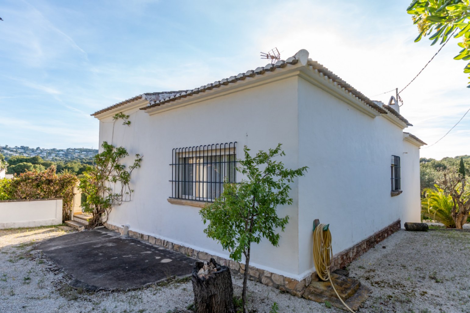 Außenfassade der Villa mit weißen Wänden, vergitterten Fenstern und Natursteinsockel, umgeben von einer Kiesfläche und mediterraner Bepflanzung.