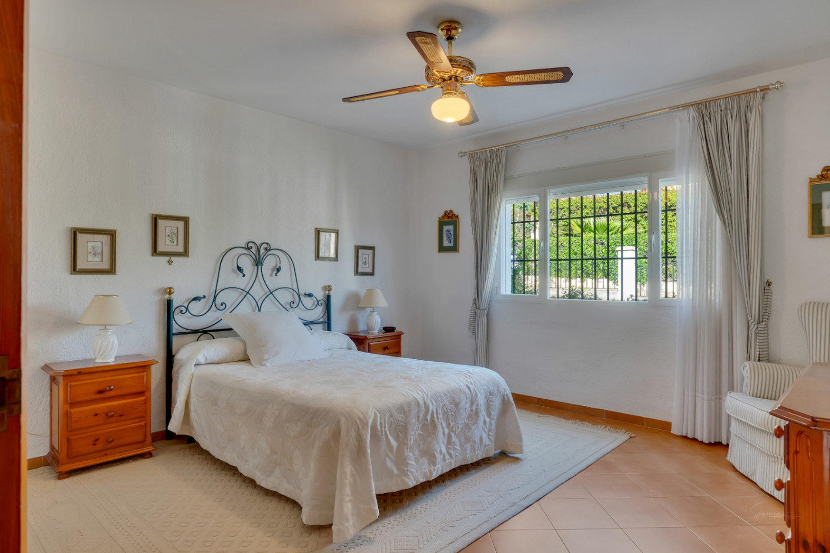 Helles Schlafzimmer mit schmiedeeisernem Kopfteil, Terrakottaboden und Deckenventilator. Das Fenster mit Sicherheitsgittern bietet natürliches Licht und Blick in den Garten.