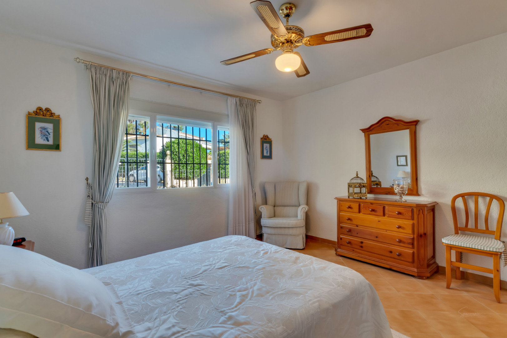 Helles Schlafzimmer mit Terrakottaboden, traditionellen Holzmöbeln, Deckenventilator und einem Fenster mit Blick in den Garten.