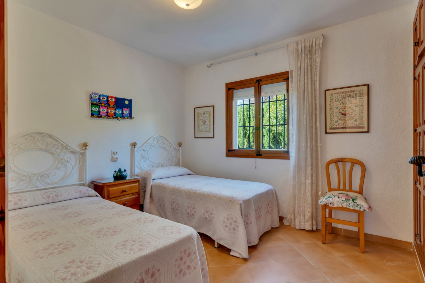 Helles Schlafzimmer mit zwei Einzelbetten, weißen schmiedeeisernen Kopfteilen, Terrakottaböden und einem klassischen Holzfenster.