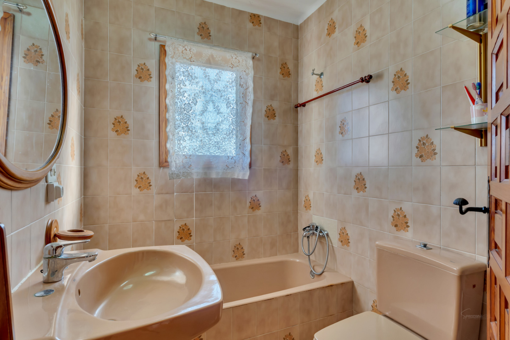 Traditionelles Badezimmer mit beige gemusterten Fliesen, passender Sanitärausstattung und Badewanne. Ein ovaler Holzspiegel und Tageslicht ergänzen das klassische Design.