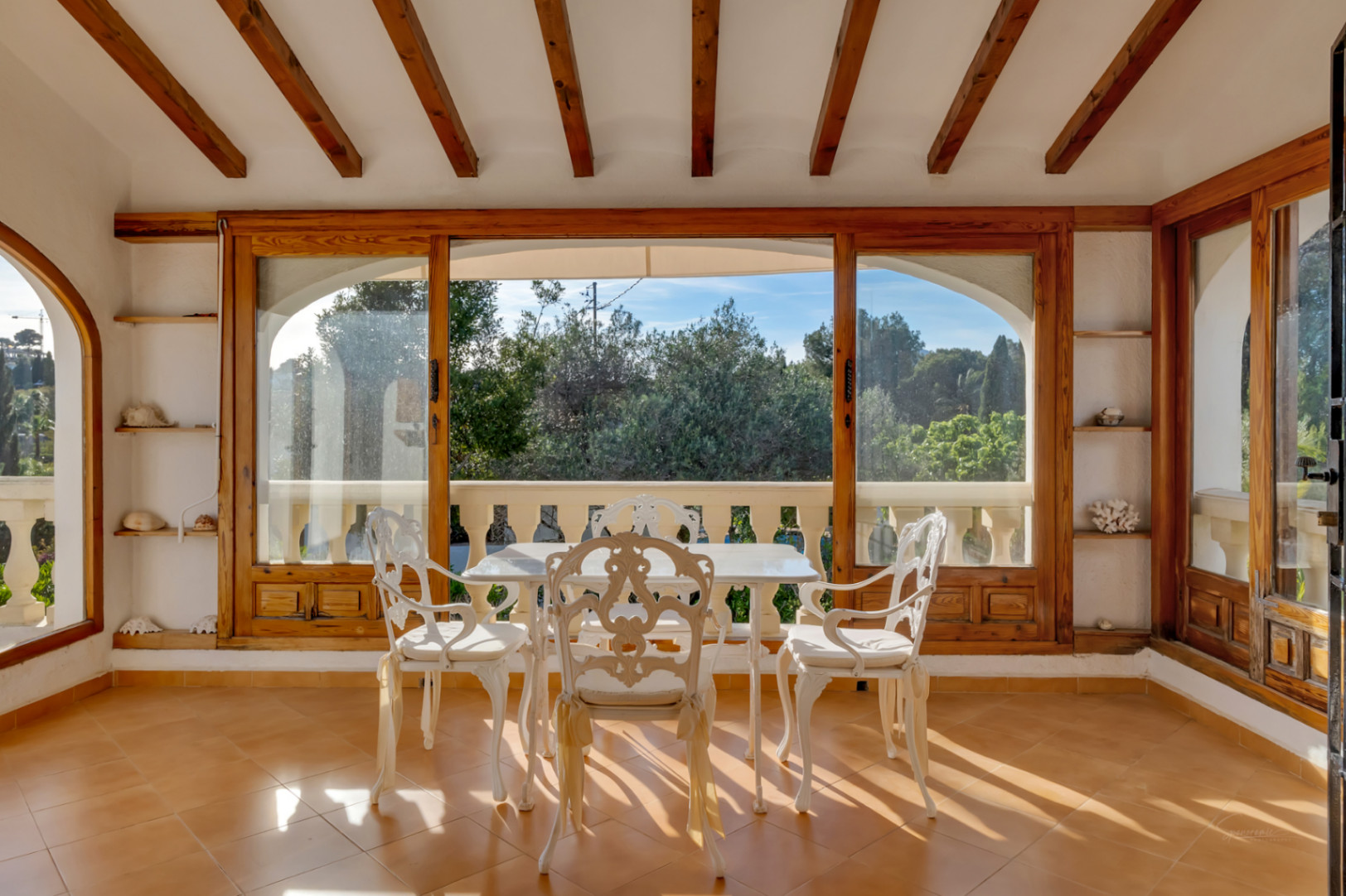 Verglaste Terrasse mit Holzbalkendecke, Terrakottaboden und weißen schmiedeeisernen Möbeln mit Blick auf den mediterranen Garten.