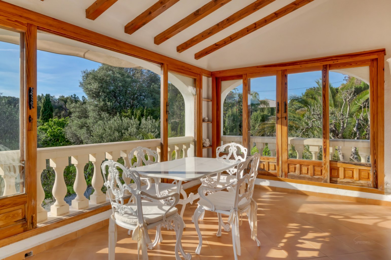 Verglaste Terrasse mit sichtbaren Holzbalken, weißen schmiedeeisernen Möbeln und großen Fenstern mit Blick auf den mediterranen Garten.