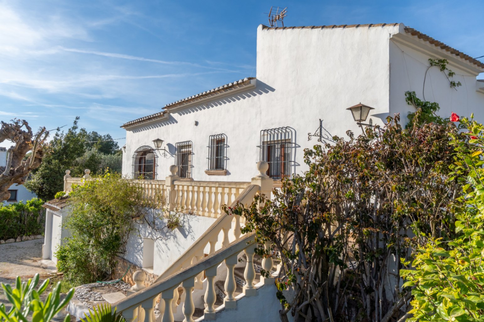 Weiße Fassade im mediterranen Stil mit Außentreppe und Balustrade, Fenstern mit schmiedeeisernen Gittern und eingewachsener Gartenbepflanzung.