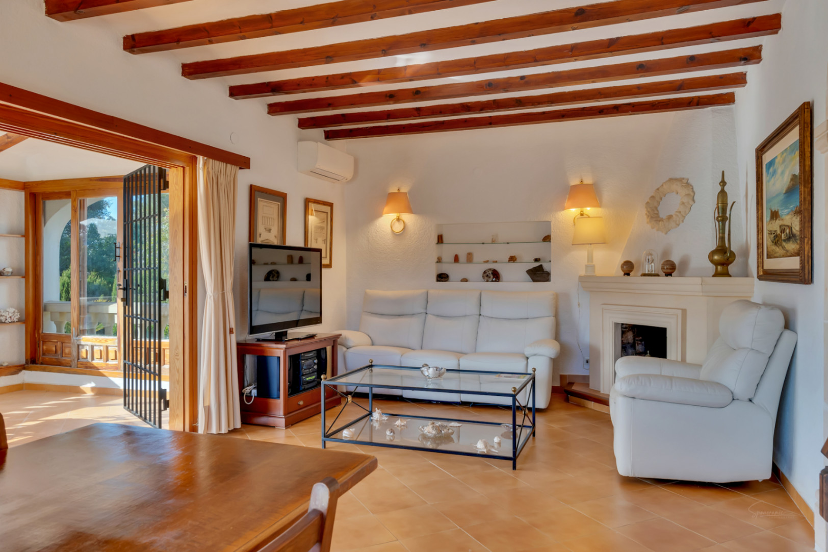 Wohnzimmer mit sichtbaren Deckenbalken aus Holz, klassischem Kamin und Terrakotta-Fliesen. Große Glastüren bieten einen direkten Zugang zur hellen Terrasse.