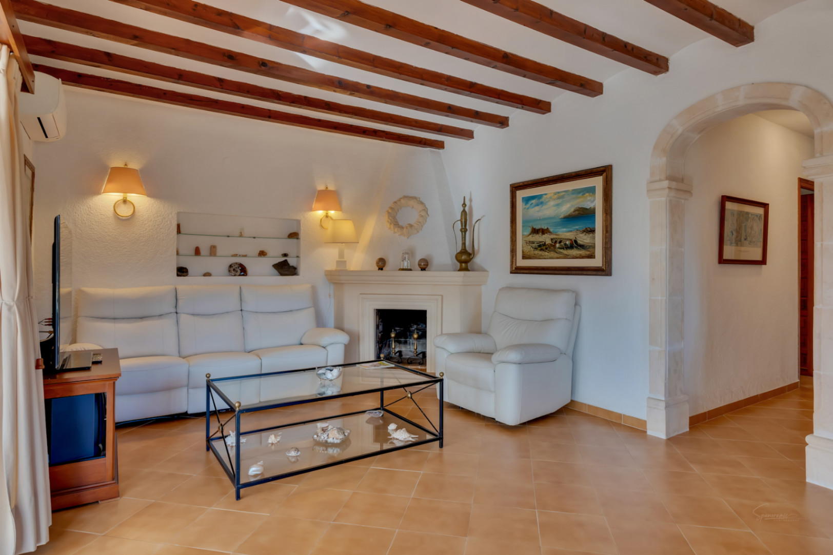Wohnzimmer mit sichtbaren Holzbalken, klassischem Kamin und einem traditionellen Rundbogen aus Tosca-Stein. Weiße Ledersofas und warme Keramikfliesen.