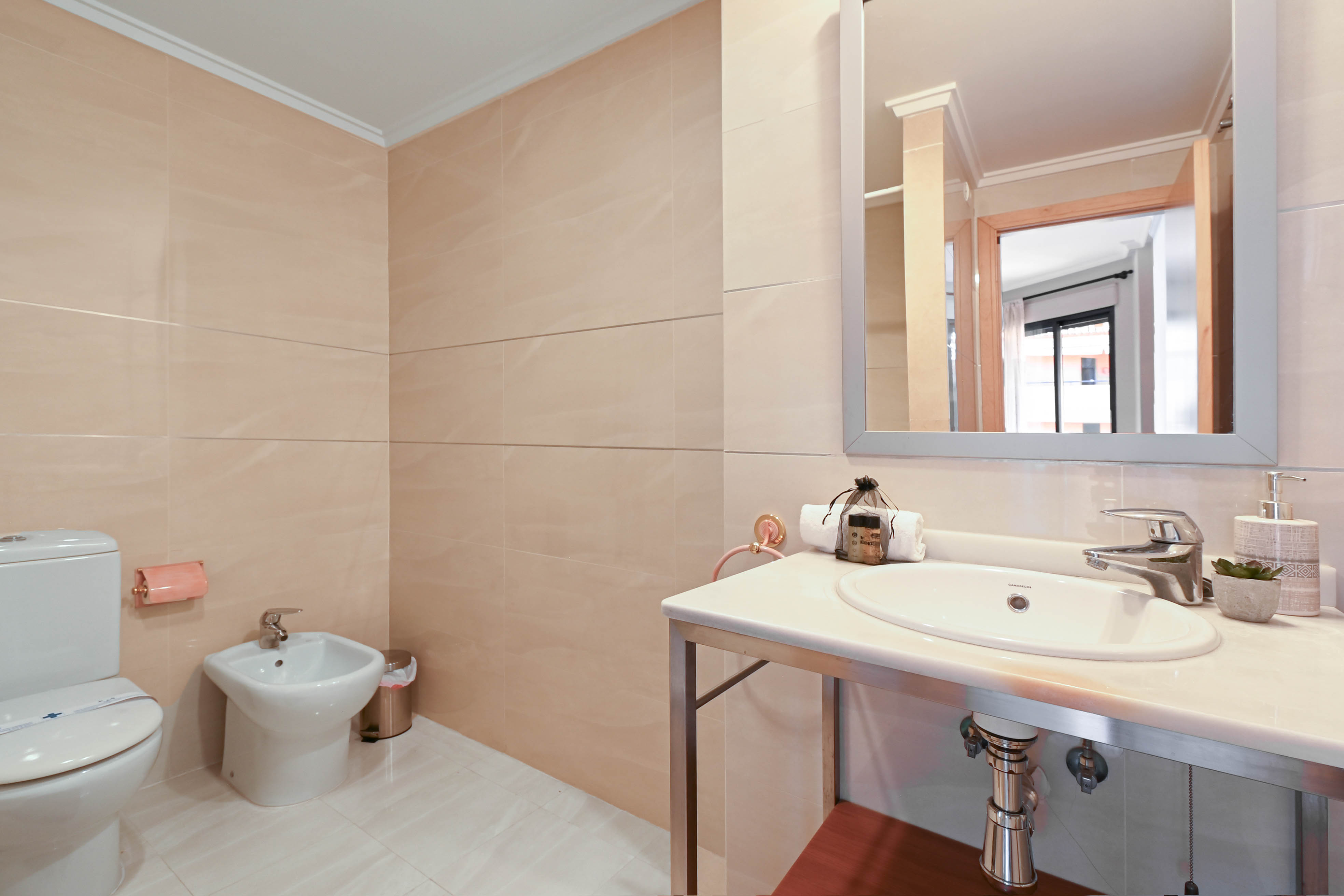 Badezimmer mit beigefarbenen Wandfliesen, ausgestattet mit Bidet, WC und einem Waschtisch aus weißem Stein auf einem modernen Chromgestell.