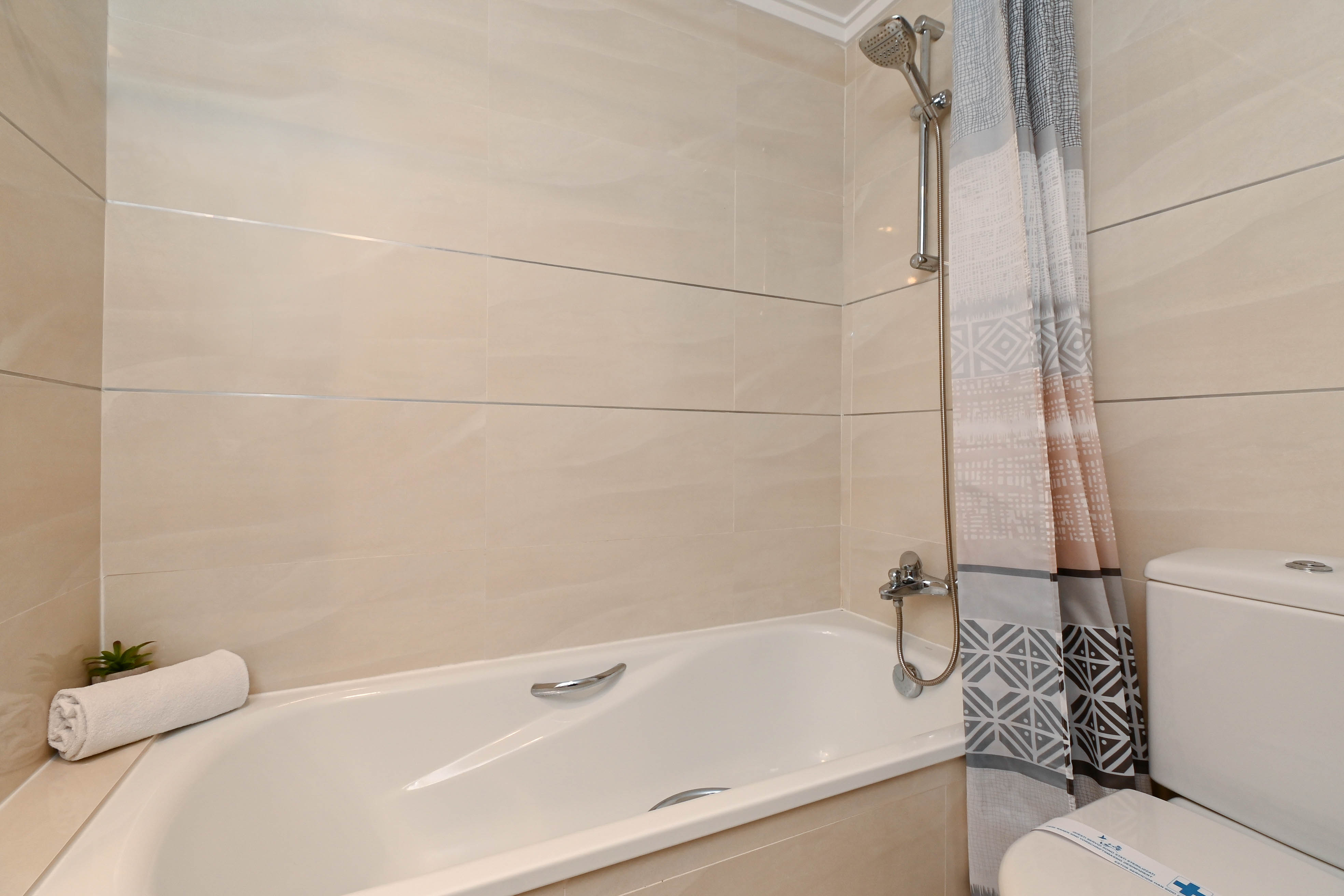 Funktionales Badezimmer mit Badewanne, verchromten Armaturen und beigen Wandfliesen mit horizontalen Akzenten sowie einem geometrisch gemusterten Duschvorhang.
