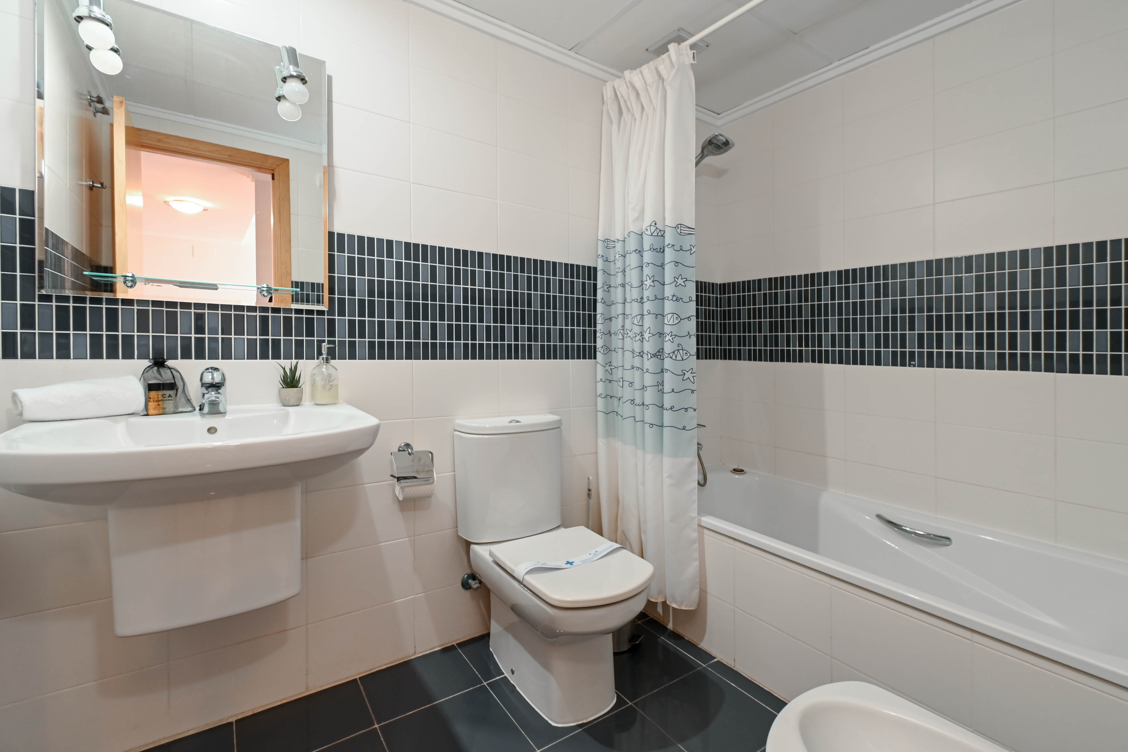 Funktionelles Badezimmer mit weißen Wandfliesen und dunkler Mosaikbordüre, ausgestattet mit Badewanne, Waschbecken und beleuchtetem Spiegel.