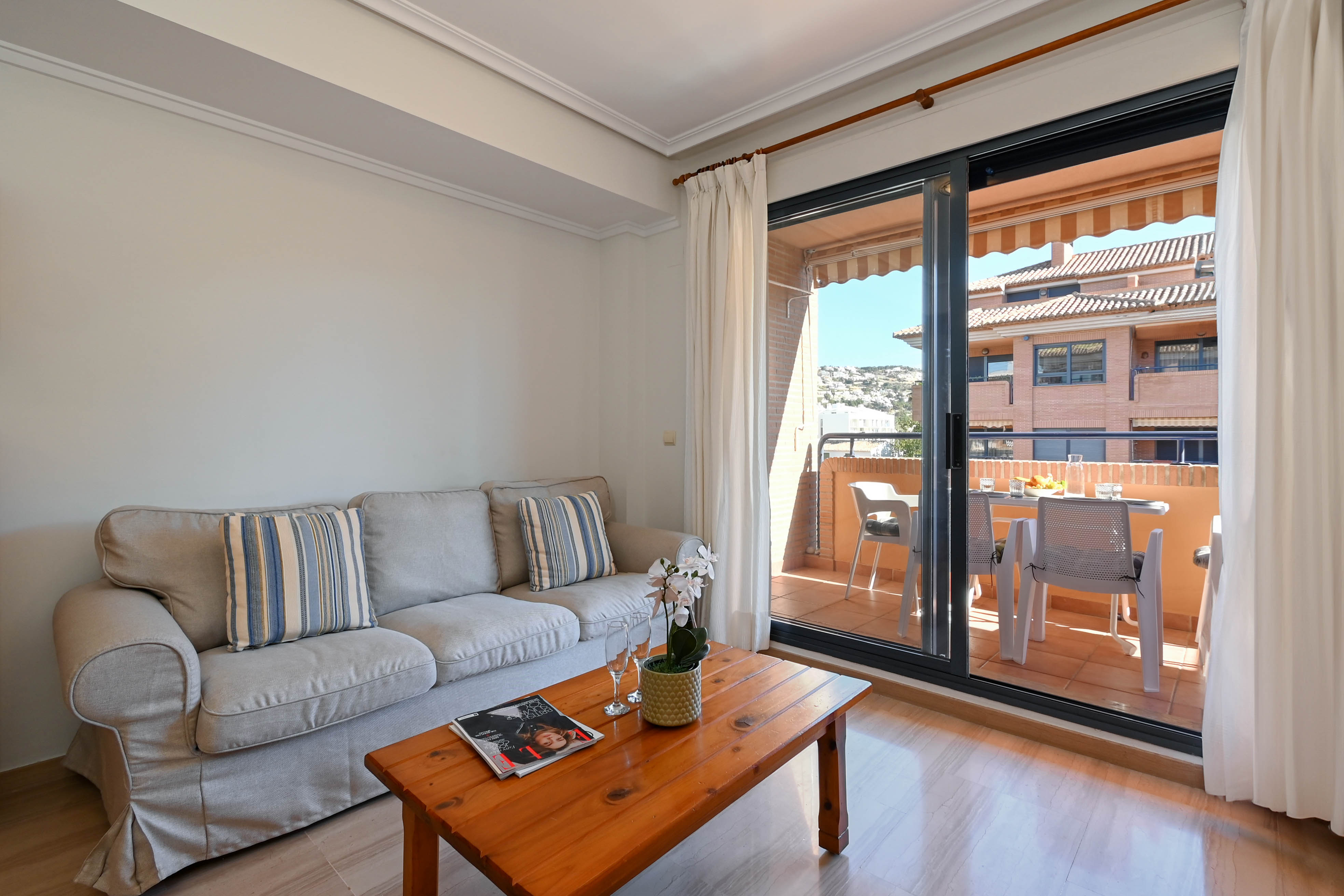 Heller Wohnbereich mit grauem Sofa und Holztisch, der durch Glasschiebetüren direkt auf einen möblierten Balkon mit Essplatz führt.