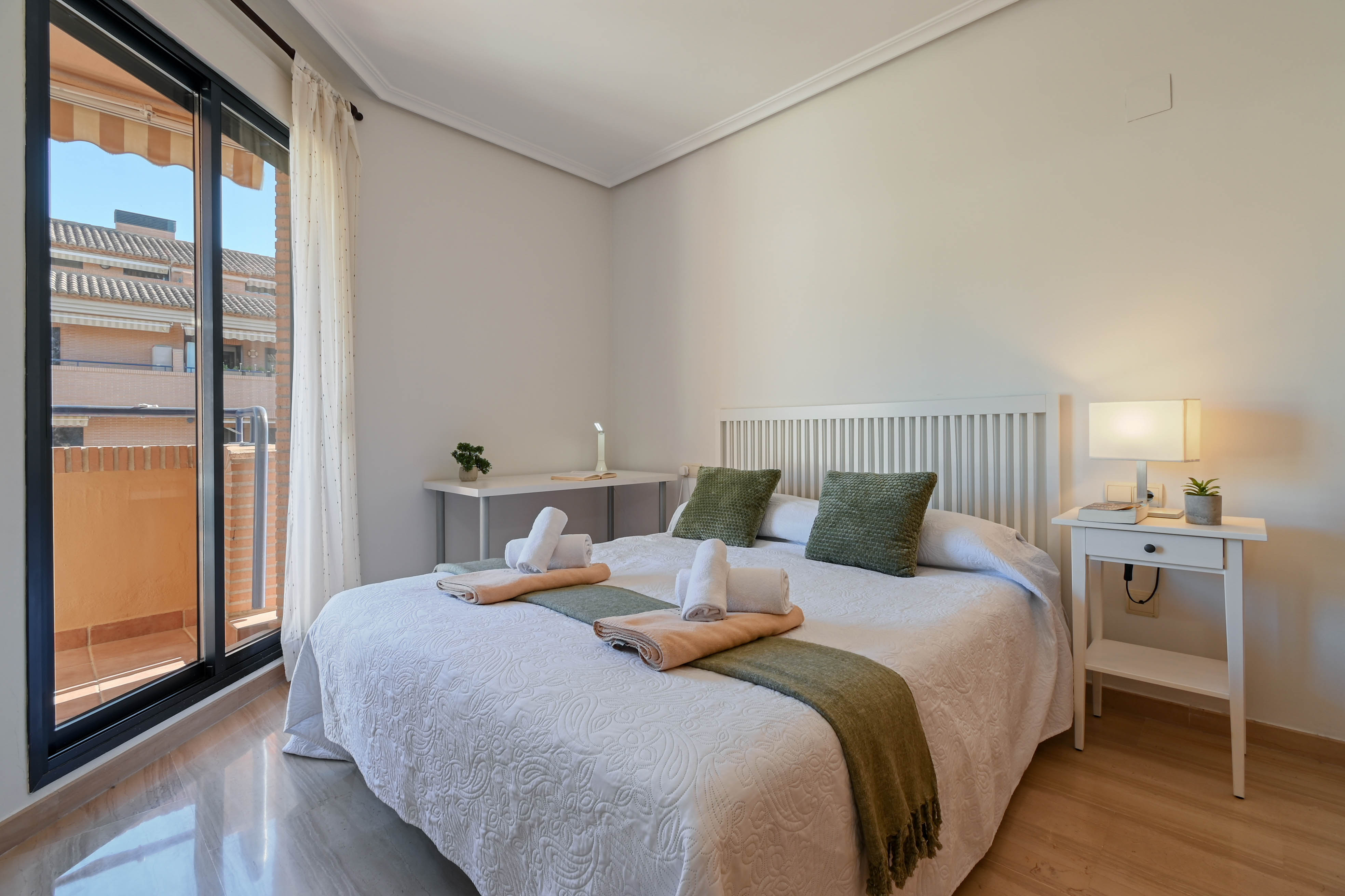 Helles Schlafzimmer mit Doppelbett, weißem Lattenkopfteil, Schreibtisch und direktem Zugang zu einem privaten Balkon durch Glasschiebetüren.