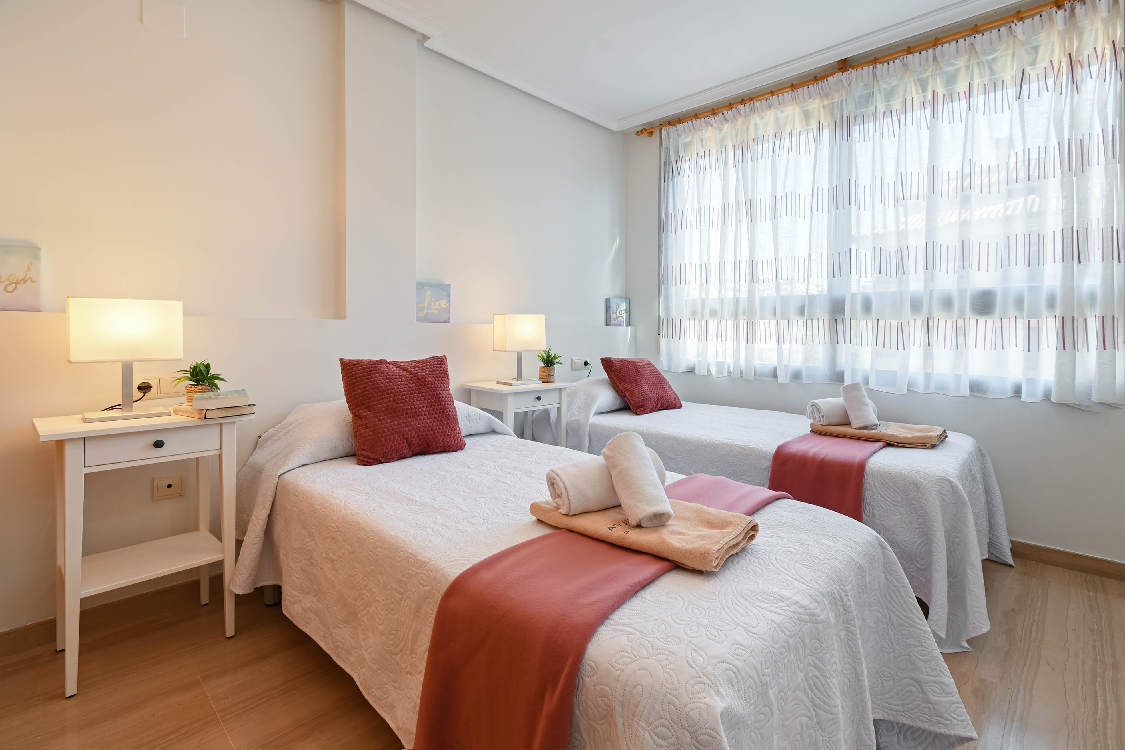 Helles Zweibettzimmer mit zwei Einzelbetten, weißen Nachttischen und hellem Holzboden. Ein großes Fenster mit gemusterten Vorhängen sorgt für angenehmes Tageslicht.