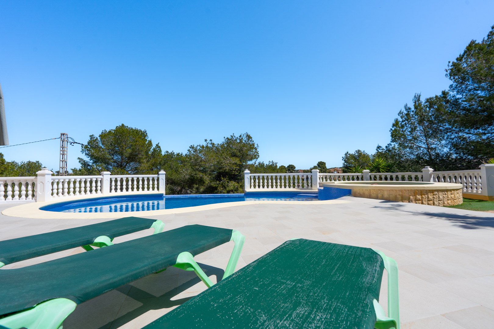 Außenanlage mit blau gefliestem Pool, weißer Balustrade und integriertem Stein-Whirlpool, umgeben von Pinien und Sonnenliegen auf hellem Steinboden.