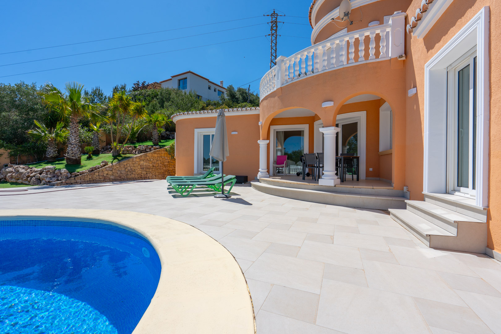 Außenbereich mit geschwungenem Pool und bogenförmiger Veranda mit weißen Säulen. Die terracottafarbene Fassade wird durch klassische Balustraden und Palmen ergänzt.